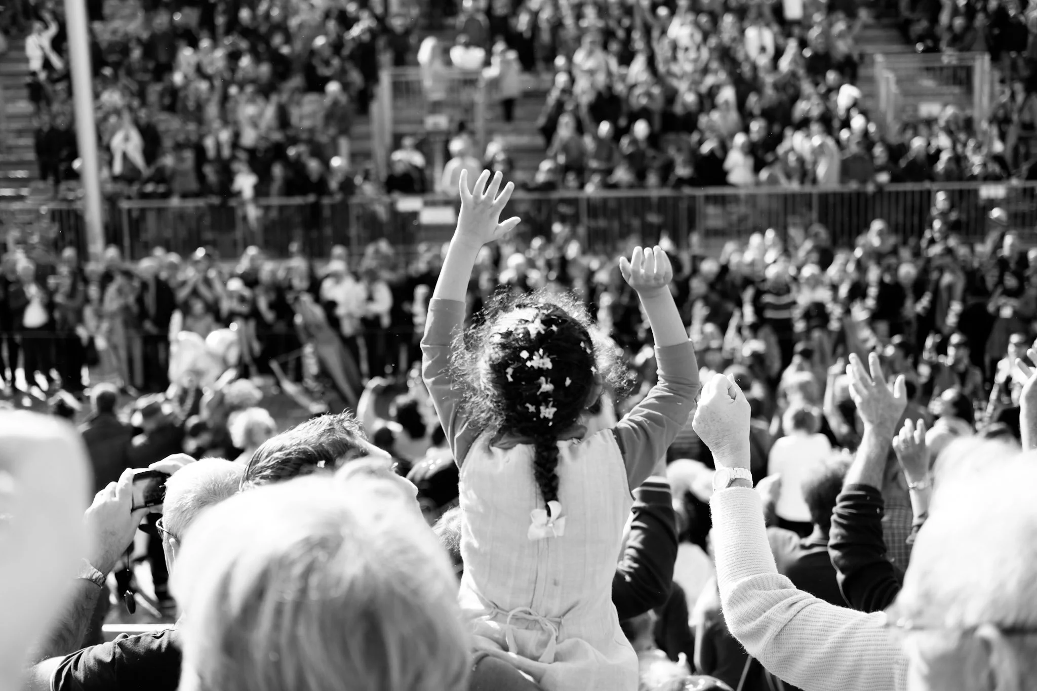 A person with curly hair raising their hand in a crowd at an outdoor event, with many other people sitting on bleachers in the background.