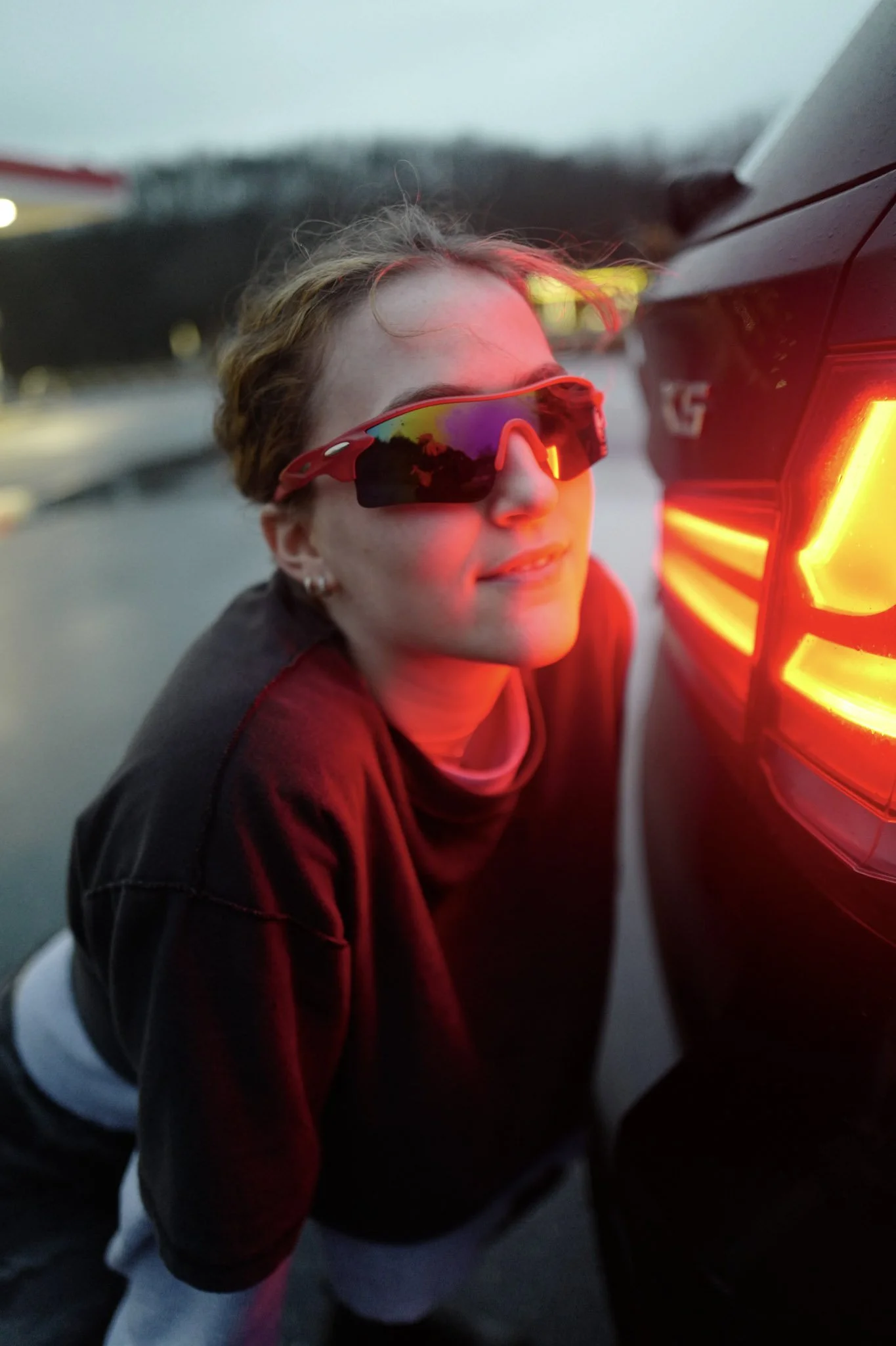 Person wearing sports sunglasses, leaning against a vehicle with illuminated red tail lights during dusk or night.