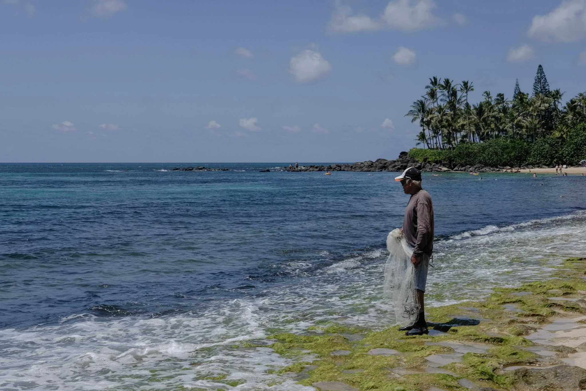 A man in a hat and sunglasses stands near the rocky shoreline with a fishing net in hand, with the ocean, a distant beach, and palm trees in the background on a bright, partly cloudy day.