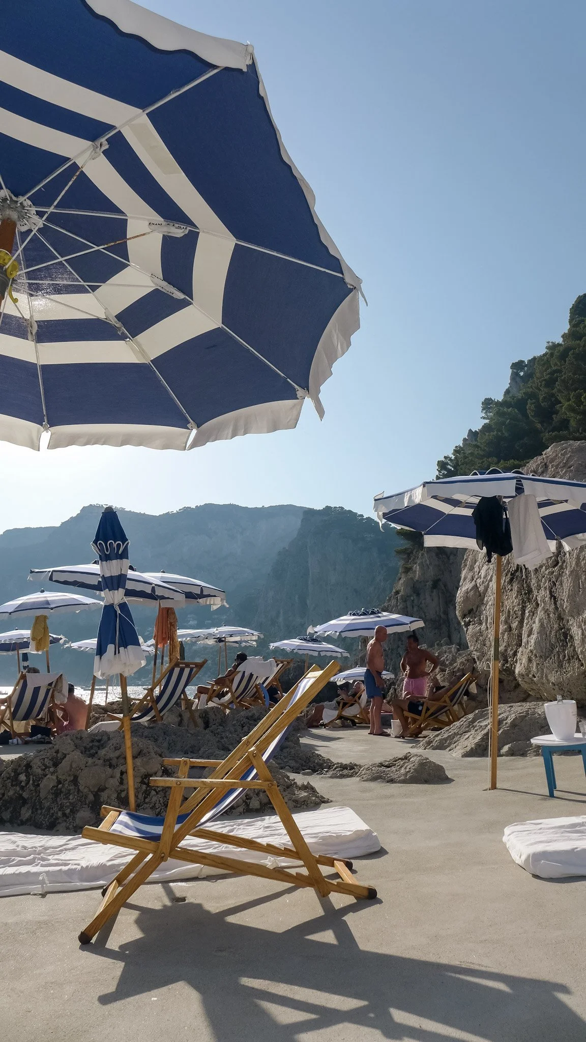 Beach scene with blue and white striped umbrellas, lounge chairs, people relaxing, rocky shoreline, and mountains in the background.