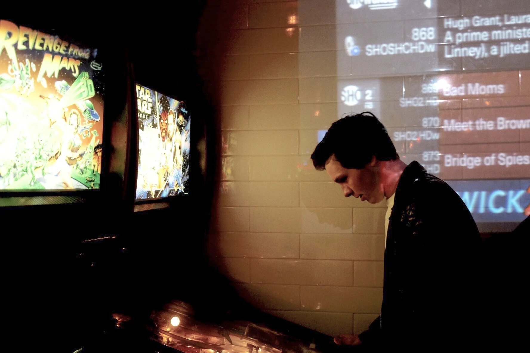 A man with dark hair looking down, standing in front of a beige tiled wall with a large screen displaying a feed of TV channel options and descriptions on his right, and movie posters featuring colorful, animated characters and scenes on his left.