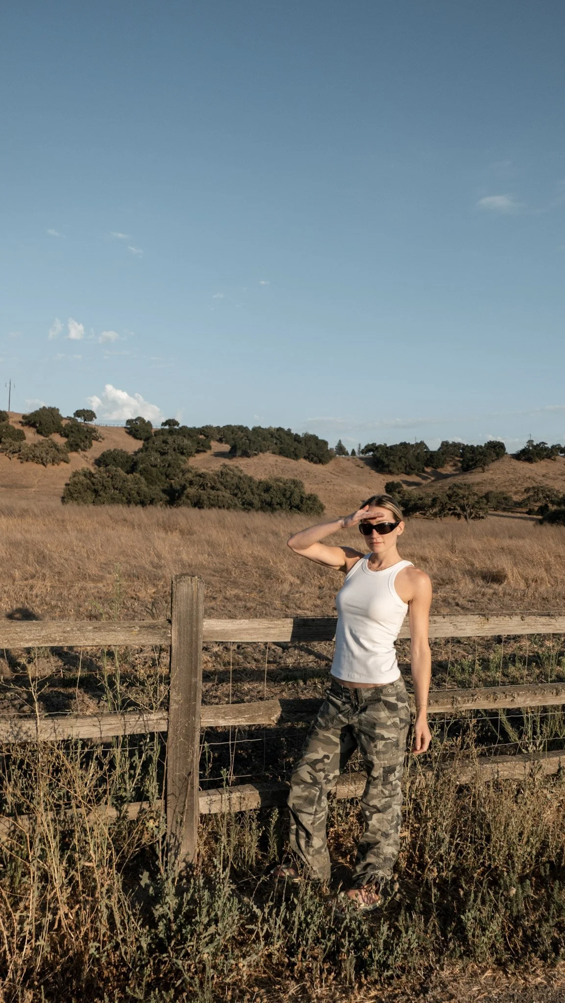 Woman in camouflage pants and white tank top standing by a wooden fence in a rural landscape, saluting the horizon with her right hand while wearing sunglasses.