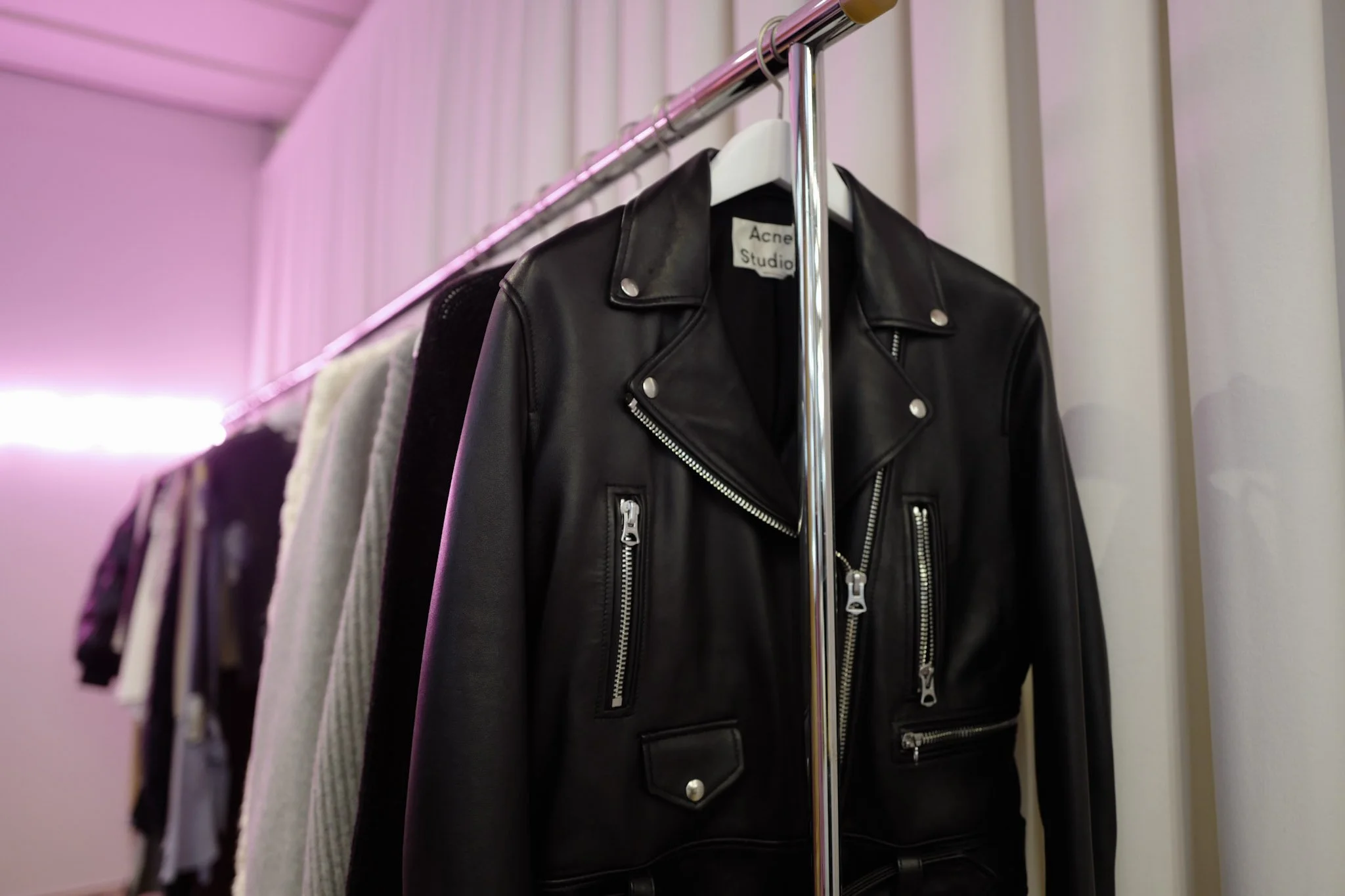 A black leather jacket hanging on a white hanger on a metal clothing rack with other garments, in a room with pastel-colored walls.