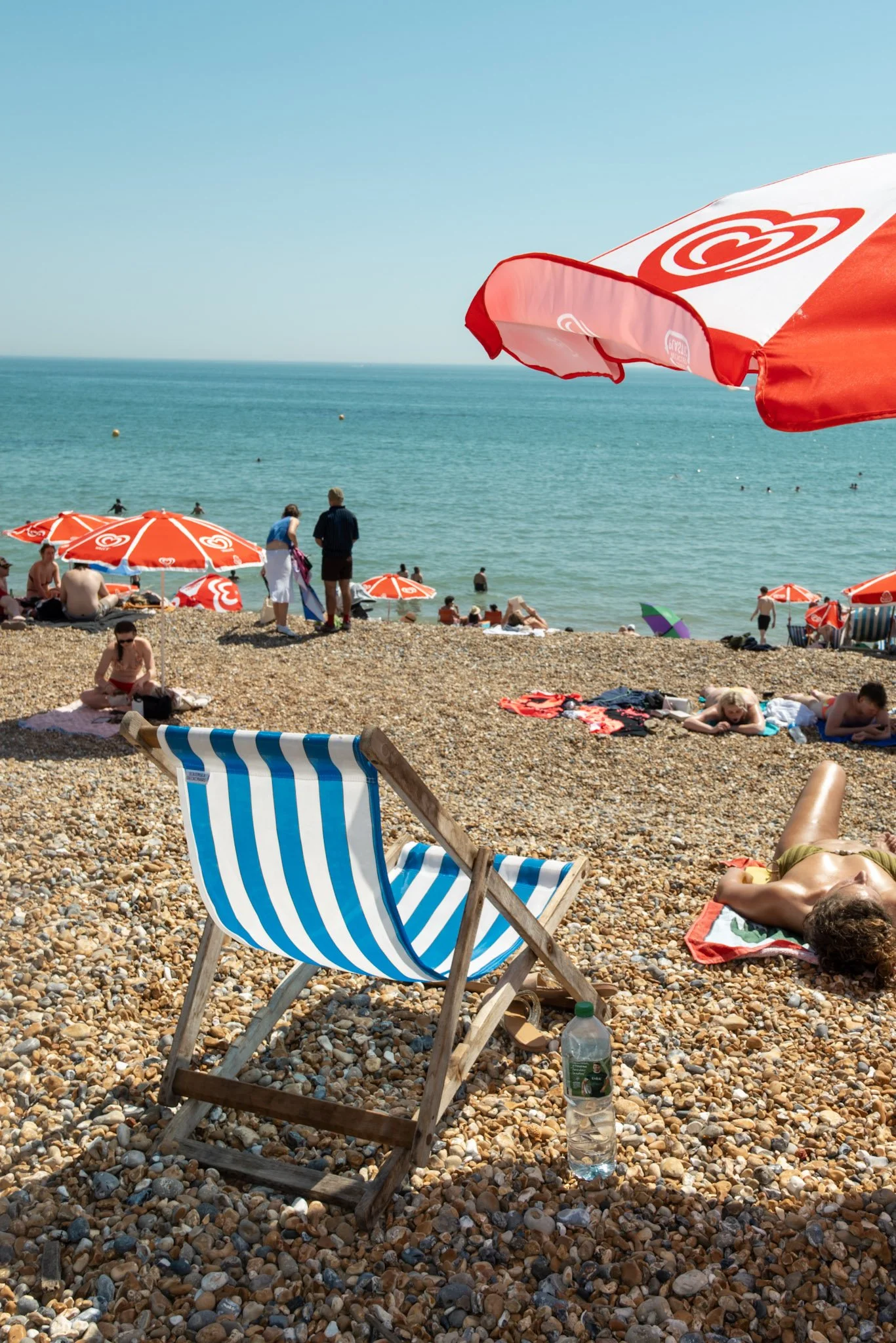 A pebble beach with sunbathers, umbrellas, and a blue ocean with a clear sky. There are people swimming and relaxing, and a striped beach chair in the foreground.