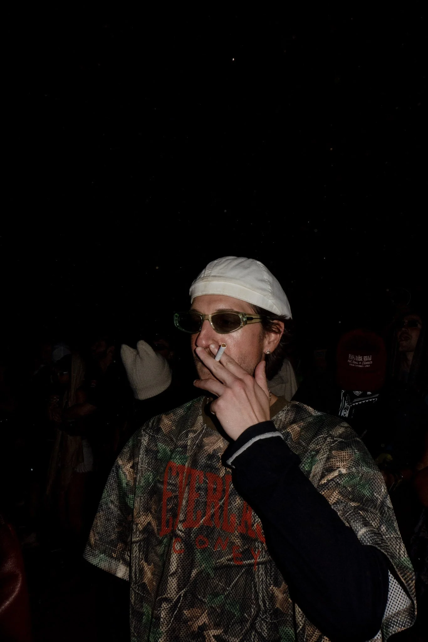 A person wearing sunglasses, a white cap, and a camouflage jacket, smoking a cigarette, in a dark setting with other people in the background.