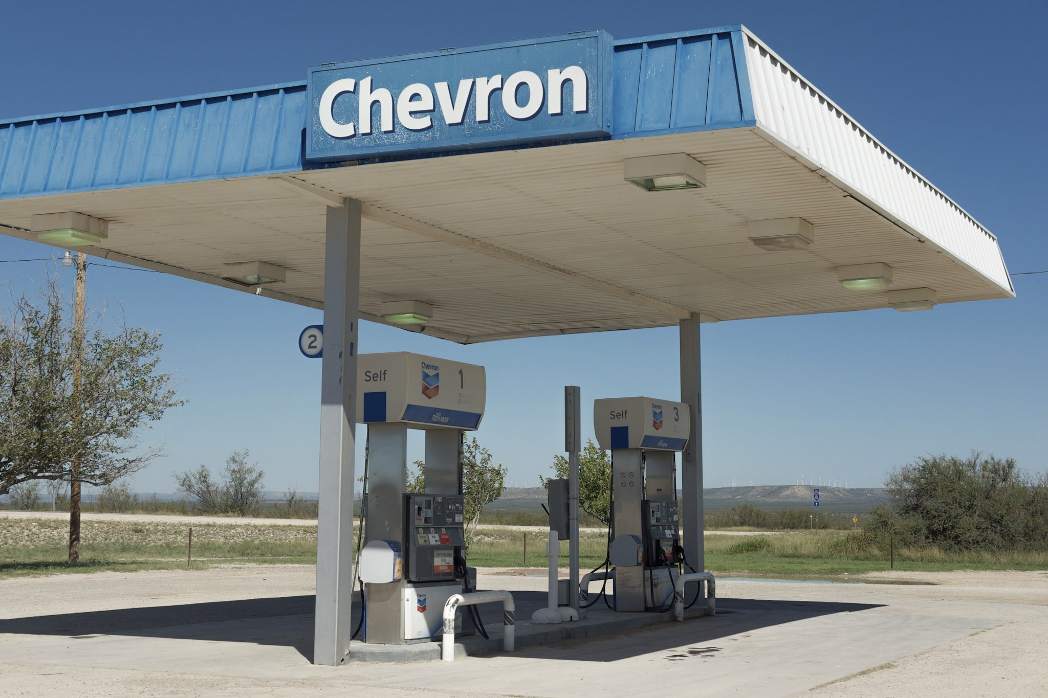 A Chevron gas station with three fuel pumps under a blue canopy, in a rural area with trees and open land in the background.