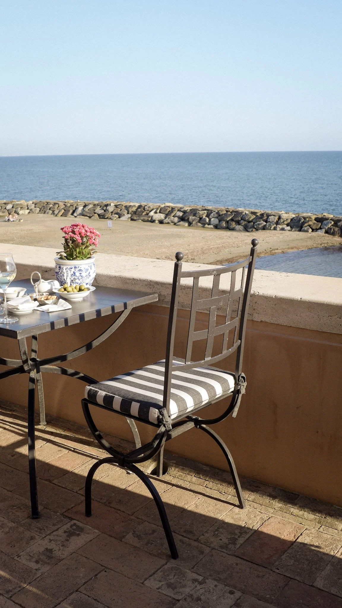 A table with a striped cushioned chair on a balcony overlooking the ocean, with a pink flower in a blue and white vase and some food and drinks on the table.