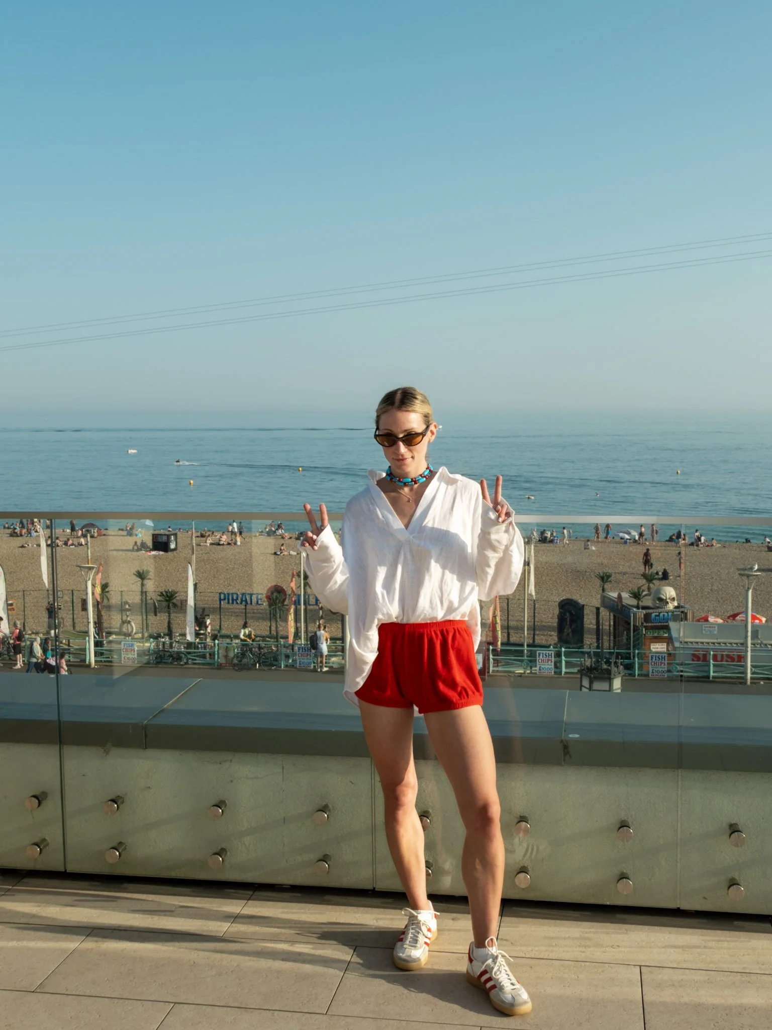 Young woman standing in front of a glass railing at a beachside, wearing sunglasses, a white long sleeve shirt, red shorts, and white sneakers with red stripes, making peace signs with both hands.