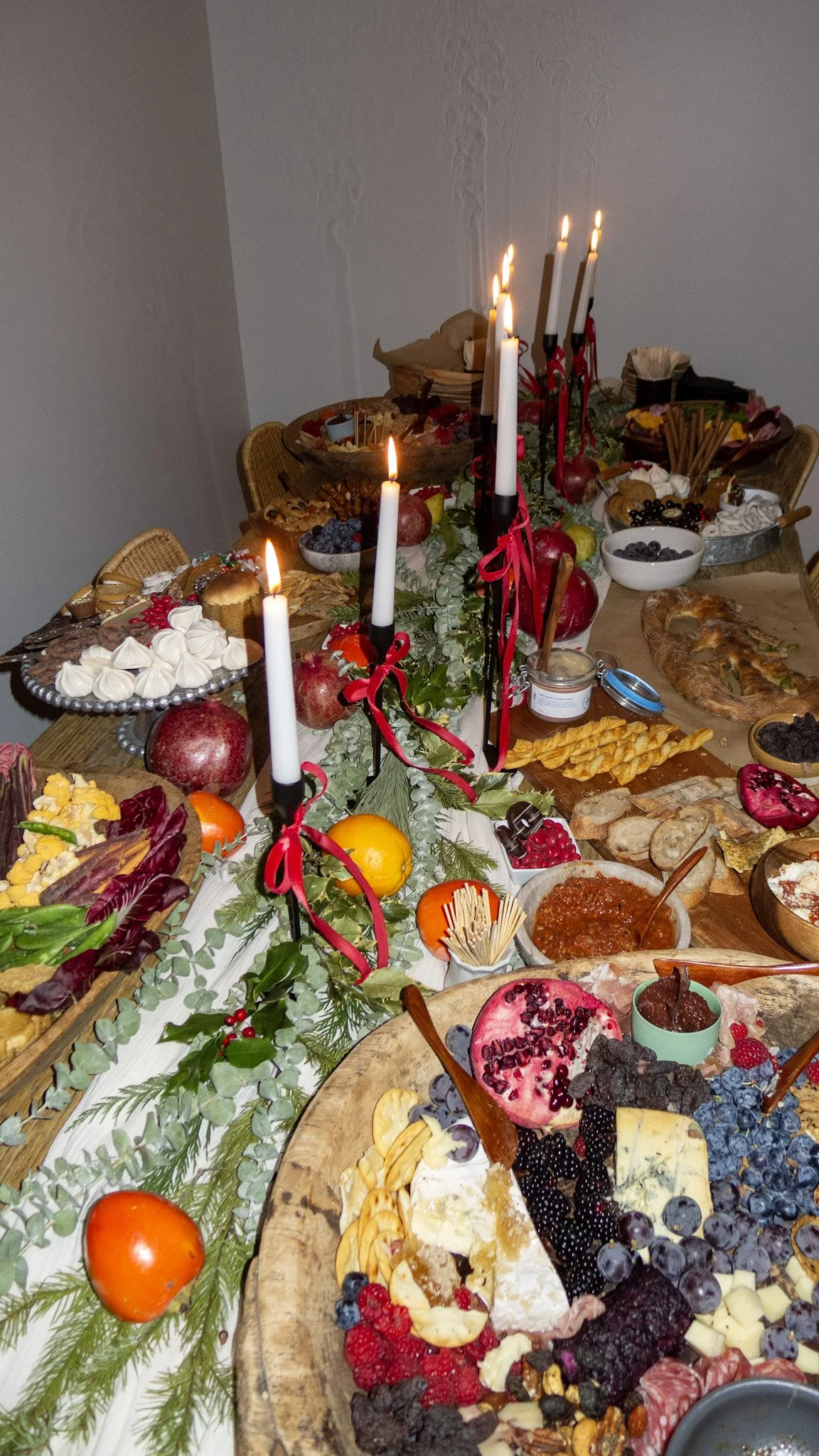 A holiday feast featuring various desserts, cheeses, fruits, and a centerpiece with candles, pomegranates, and greenery on a large wooden table.