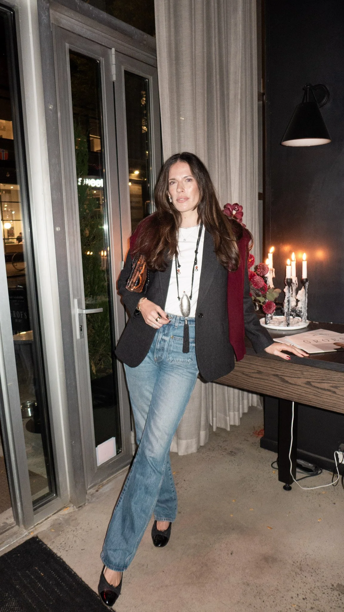A woman with long brown hair wearing a black blazer, white shirt, and high-waisted jeans standing inside a modern space with candles on a table and a decorative flower arrangement. She is looking at the camera with her left hand resting on the table 