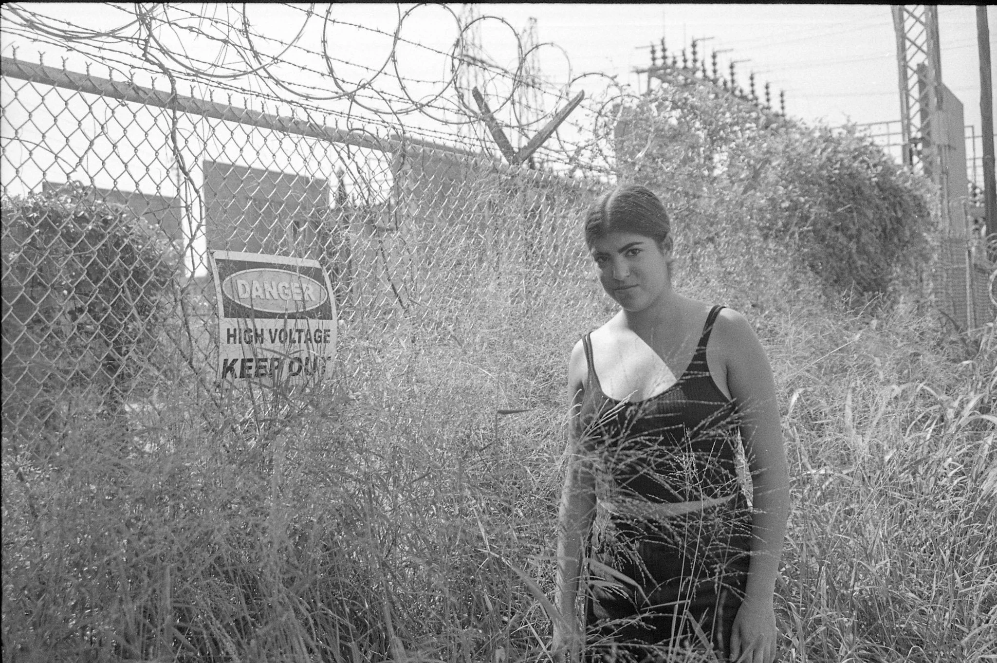 Black and white photo of a young woman standing outdoors next to a barbed wire fence with high voltage warning sign, bushes, and power lines in the background.