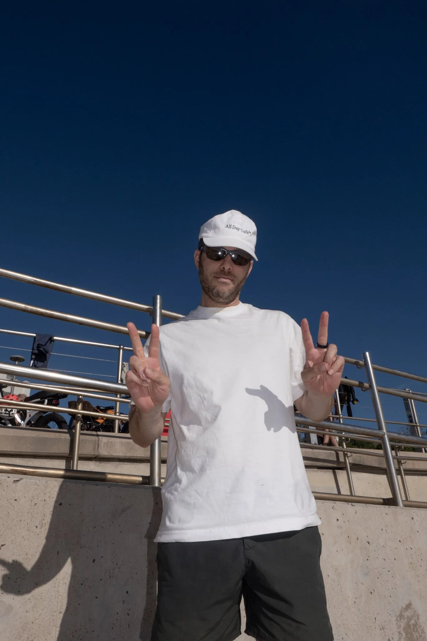 A man wearing sunglasses, a white baseball cap, and a white T-shirt making peace signs with both hands standing outdoors in front of a metal railing, with bicycles in the background and a clear blue sky above.