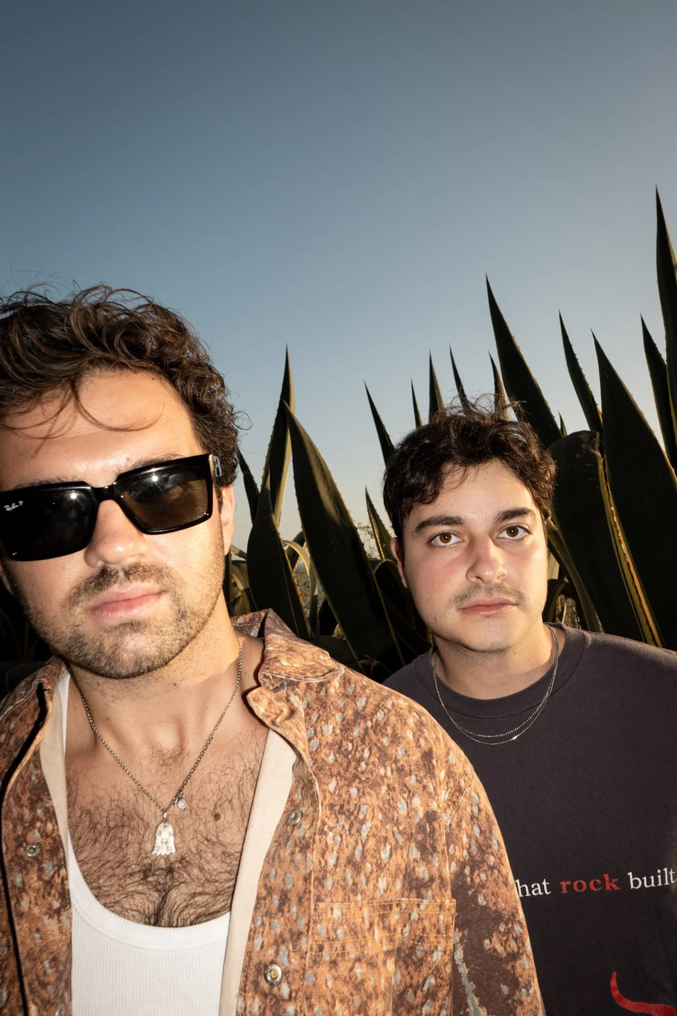 Close-up of two young men outdoors, one wearing sunglasses and a patterned shirt, the other wearing a black T-shirt, with tall plants and a clear sky in the background.