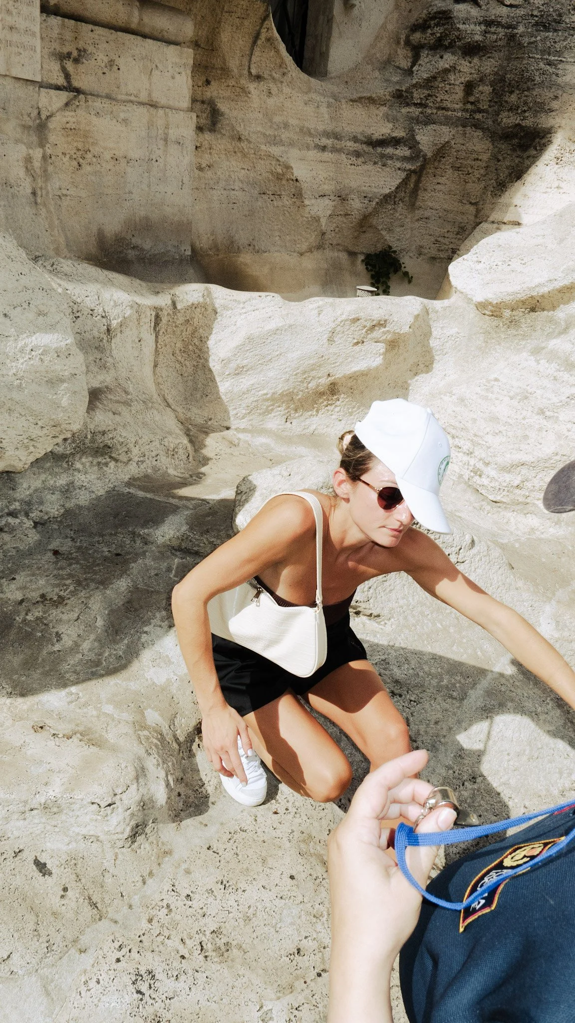 A woman crouching on sandy ground near rocky terrain, wearing sunglasses, a white cap, and a white handbag, while a person in a uniform is taking a photo of her with a camera.