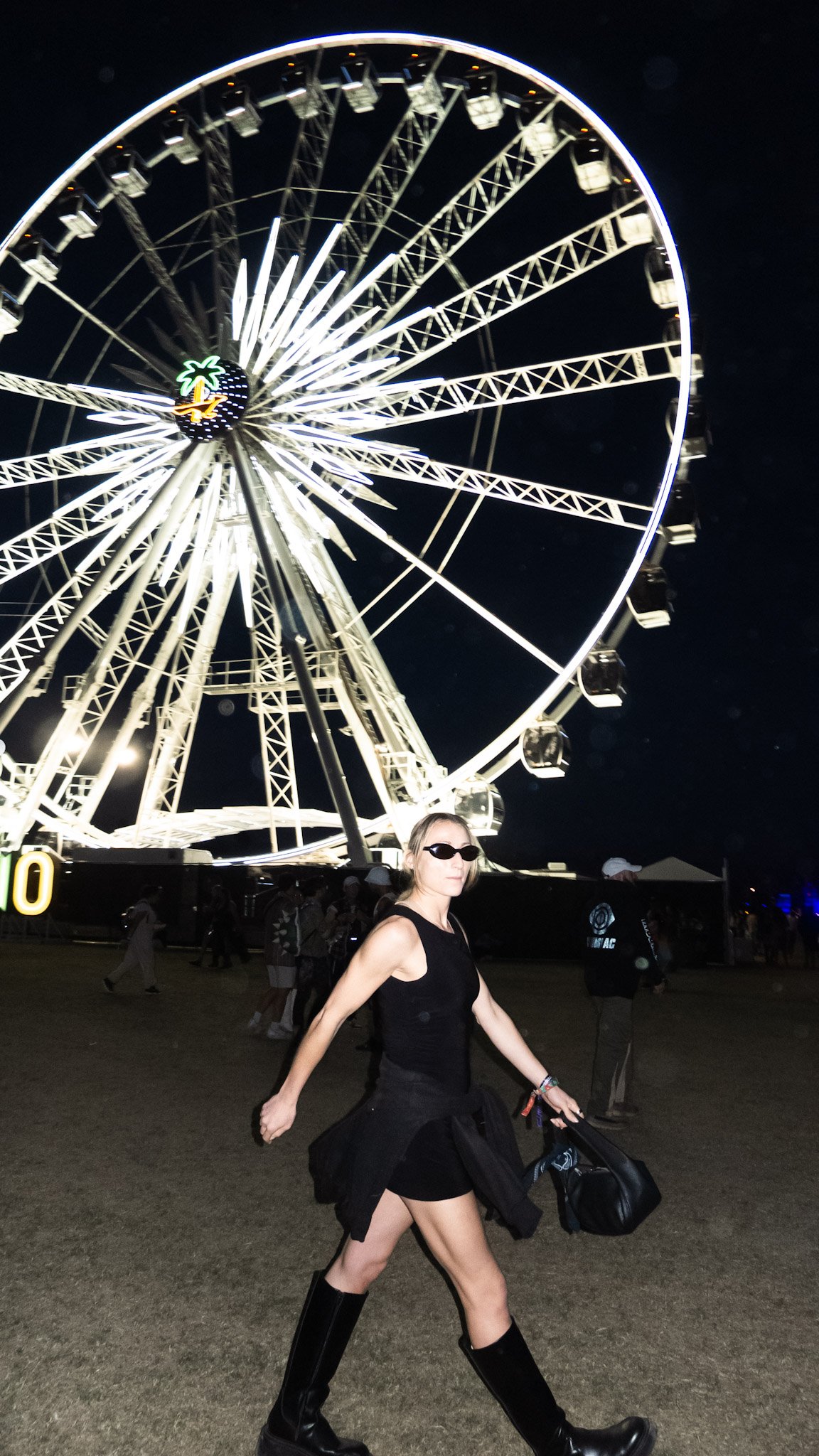 A woman in black clothing and sunglasses walking past a brightly lit Ferris wheel at night during a fair or carnival.
