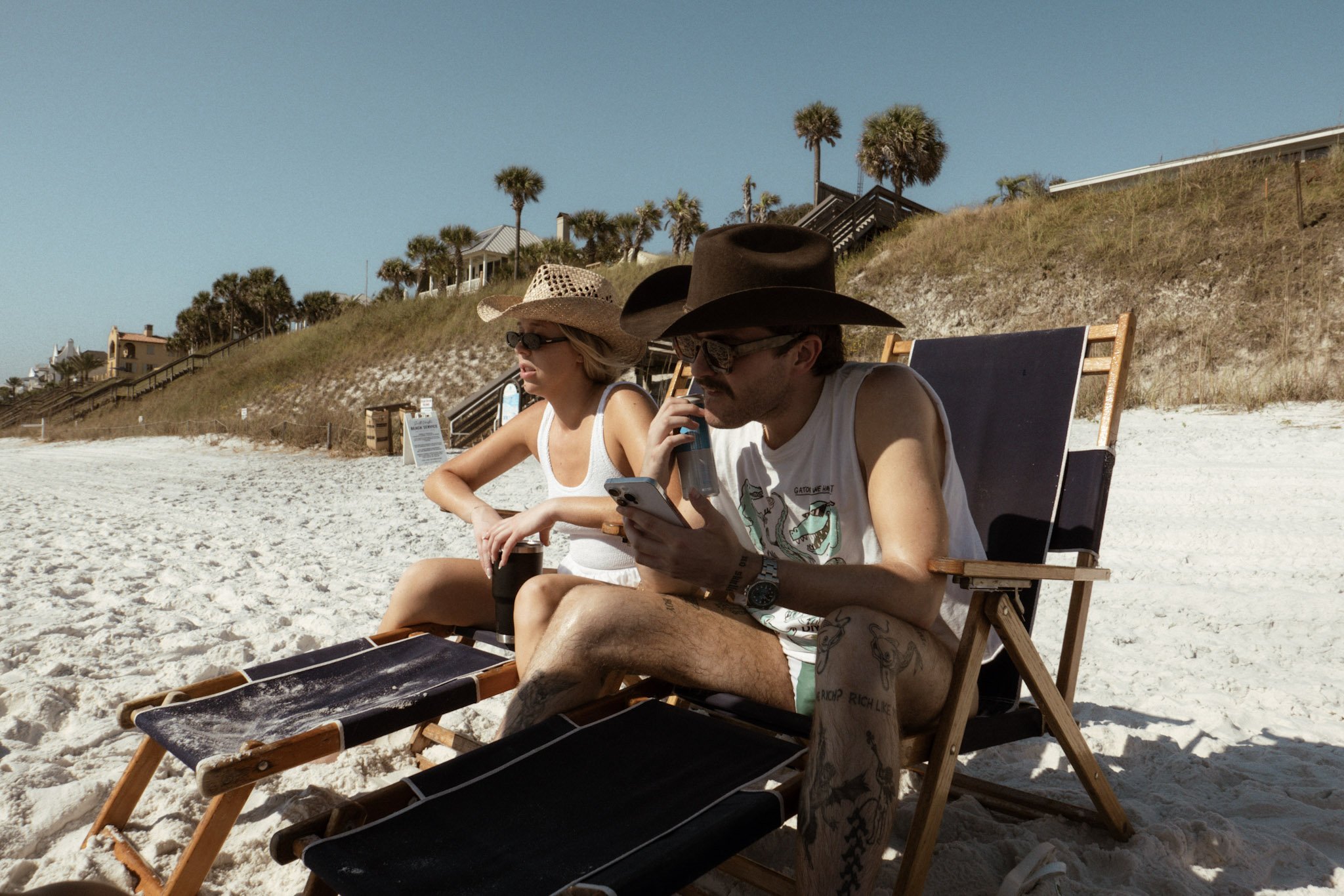 Two people sitting on beach chairs on sand, wearing hats and sunglasses. One person is wearing a tank top, and the other is wearing a sleeveless shirt. They appear to be relaxing and looking at a phone.