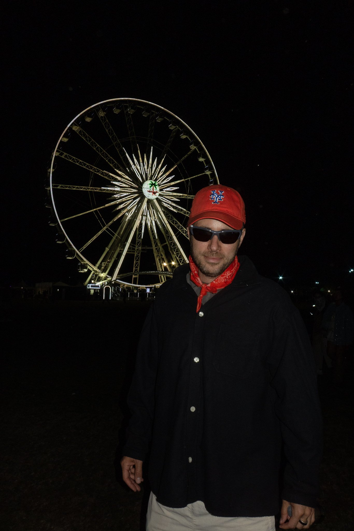 A man wearing sunglasses, a red cap with a baseball logo, and a bandana around his neck takes a selfie in front of a large illuminated Ferris wheel at night. The Ferris wheel features bright lights and a palm tree logo at its center.