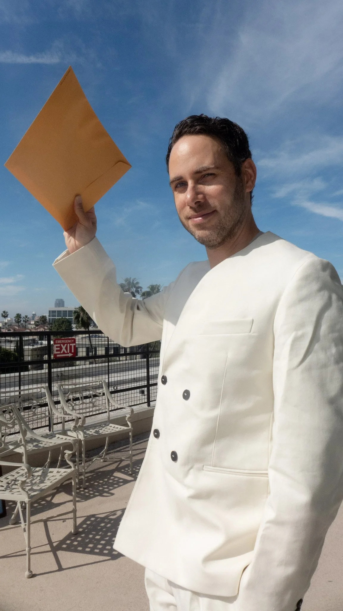 A man in a white chef's coat holding a yellow envelope on a rooftop with blue skies and city buildings in the background.