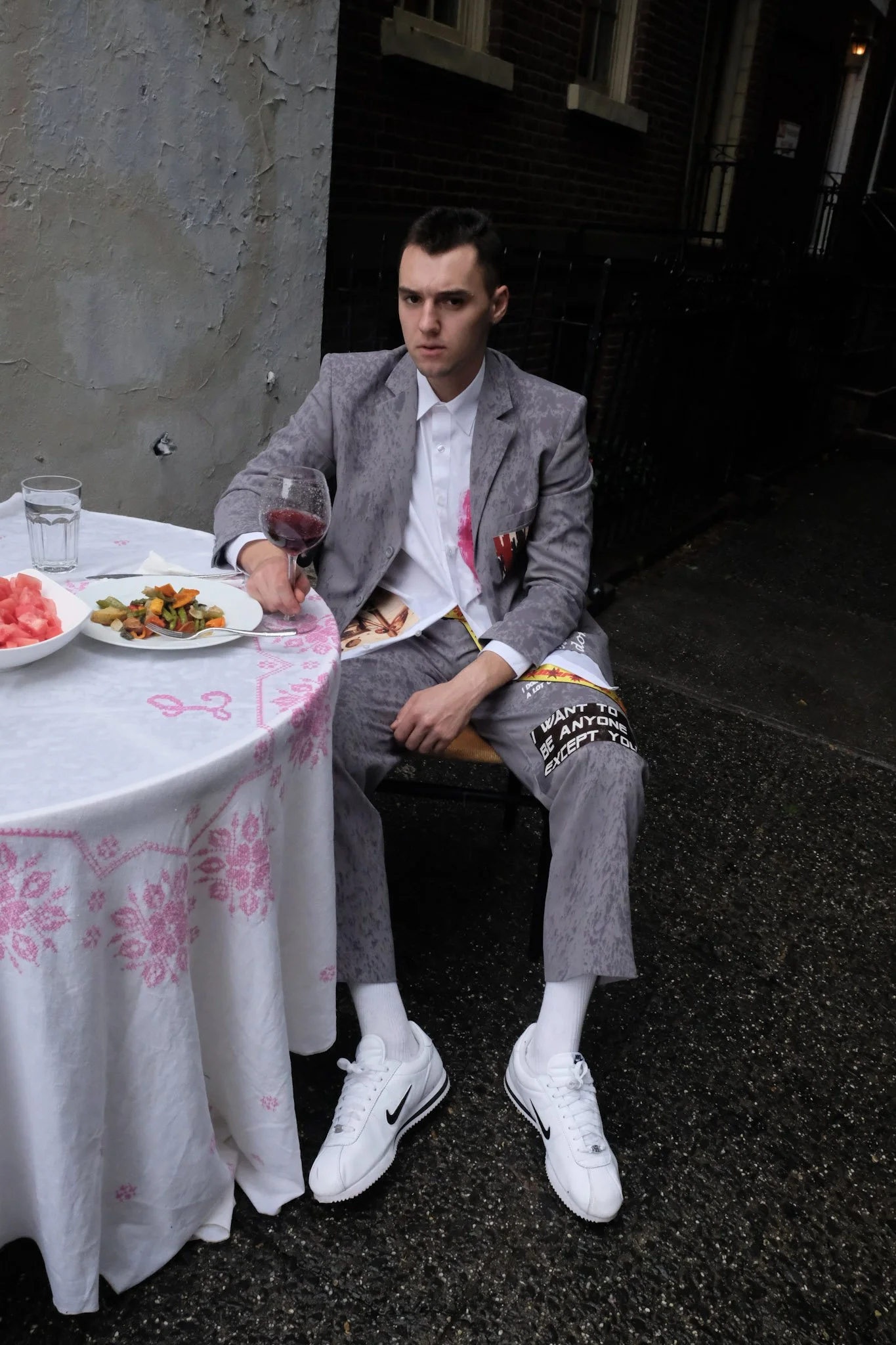 A young man sitting at an outdoor table, holding a glass of red wine, dressed in a gray suit with a white shirt, white sneakers, and baggy gray pants with black and white lettering, in a dimly lit street setting with food on the table.