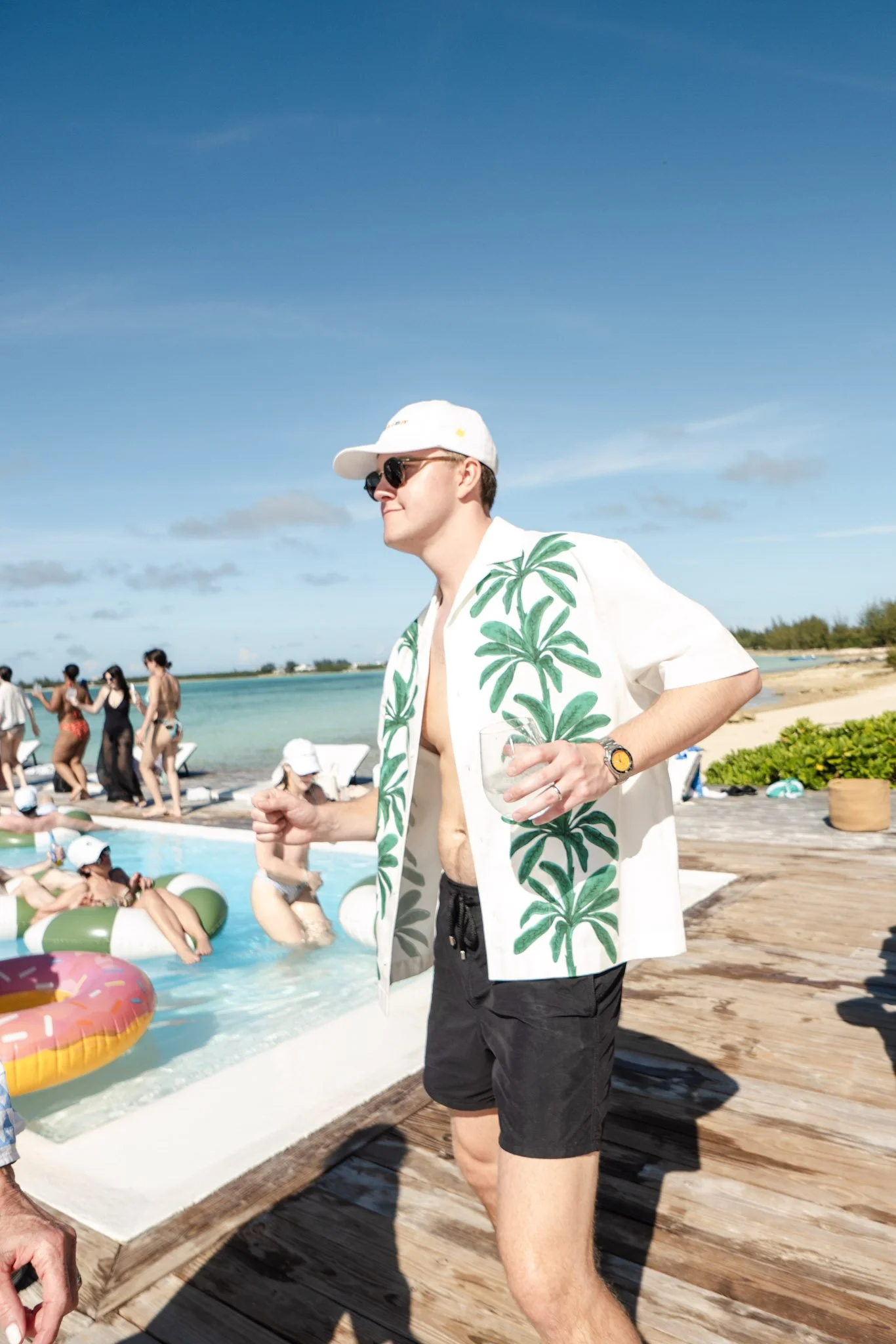 A man in a white shirt with green palm tree patterns, black shorts, sunglasses, a white cap, holding a drink, standing at a pool party on a beach with people swimming and lounging by the pool, sunny weather, blue sky.