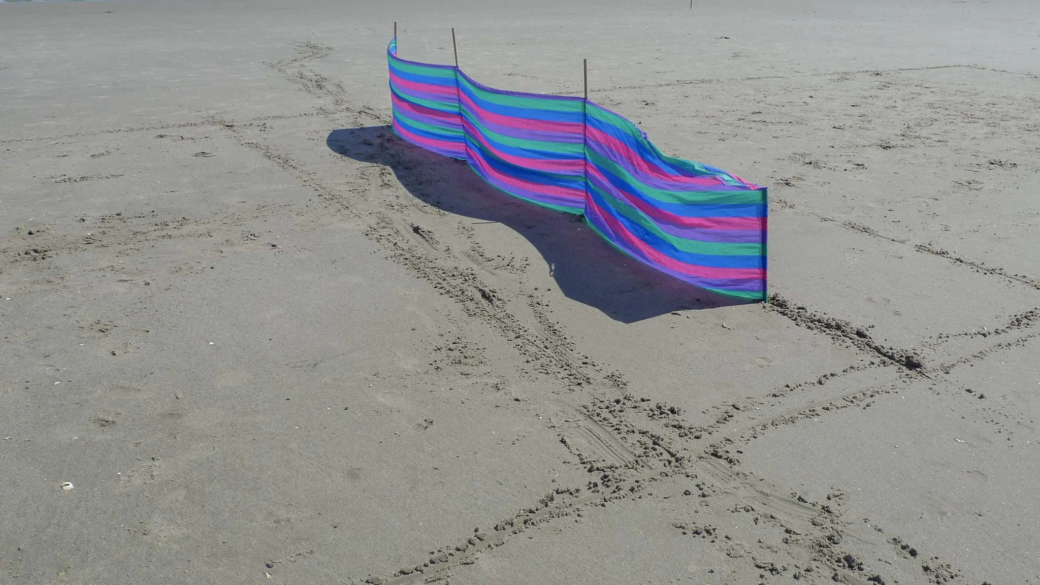Colorful striped fabric or barrier set up on a sandy beach with tire tracks and footprints, casting a shadow.
