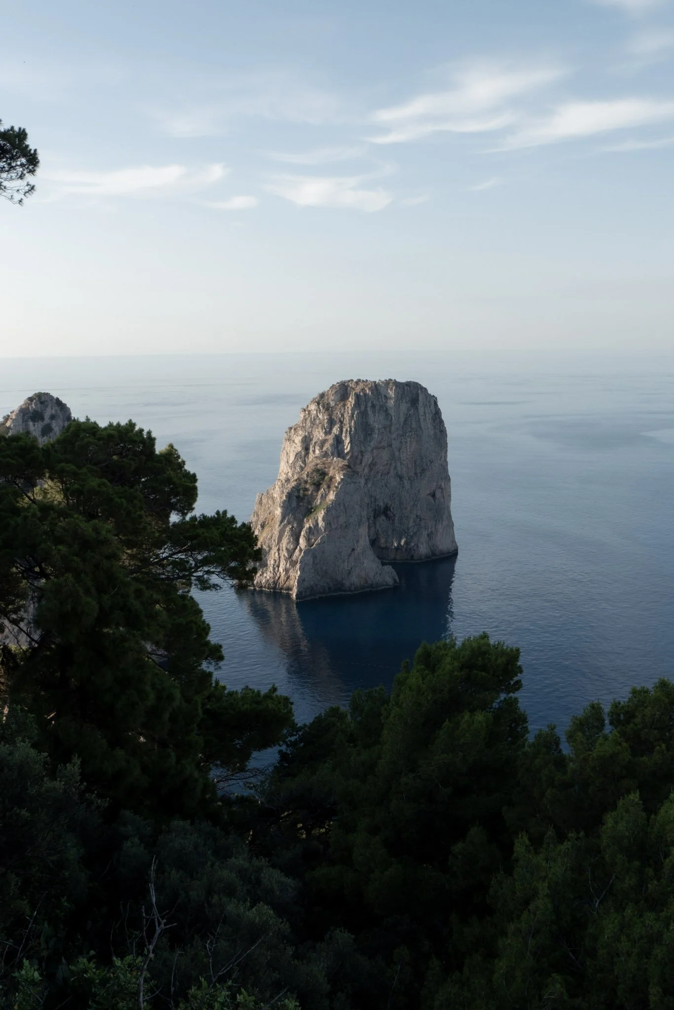 A large rock formation surrounded by water, with green trees in the foreground and a partly cloudy sky overhead.