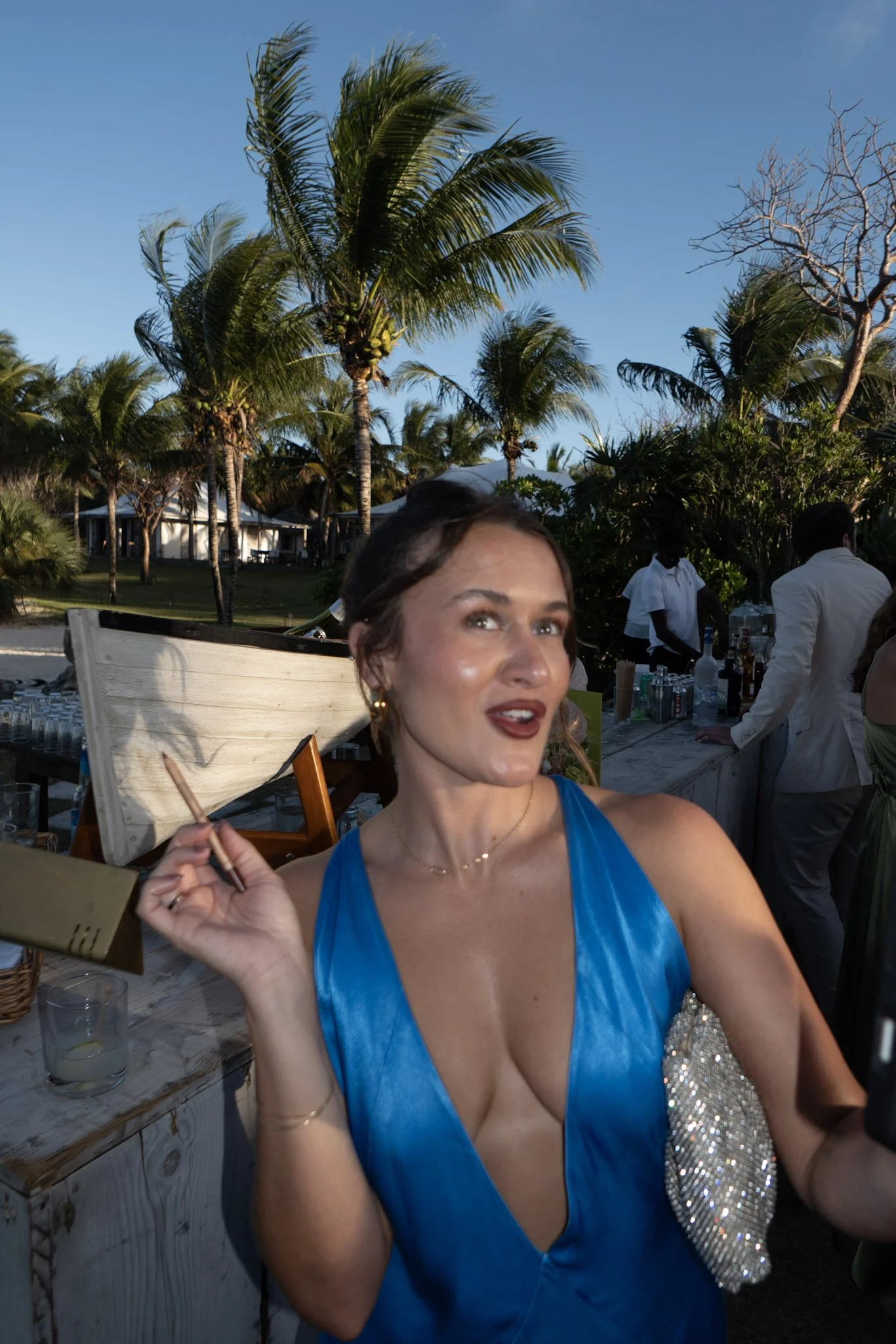 A woman wearing a blue dress holding a cigarette at an outdoor event with palm trees, a grassy area, and servers in white uniforms in the background.