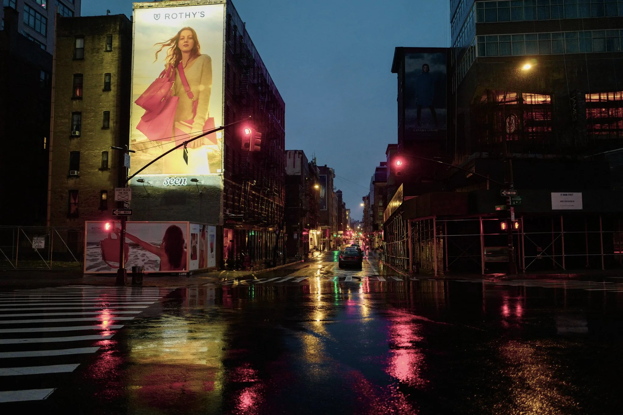City street scene at dusk with illuminated billboards, wet pavement reflecting lights, and traffic signals showing red. Buildings and advertisements are visible.