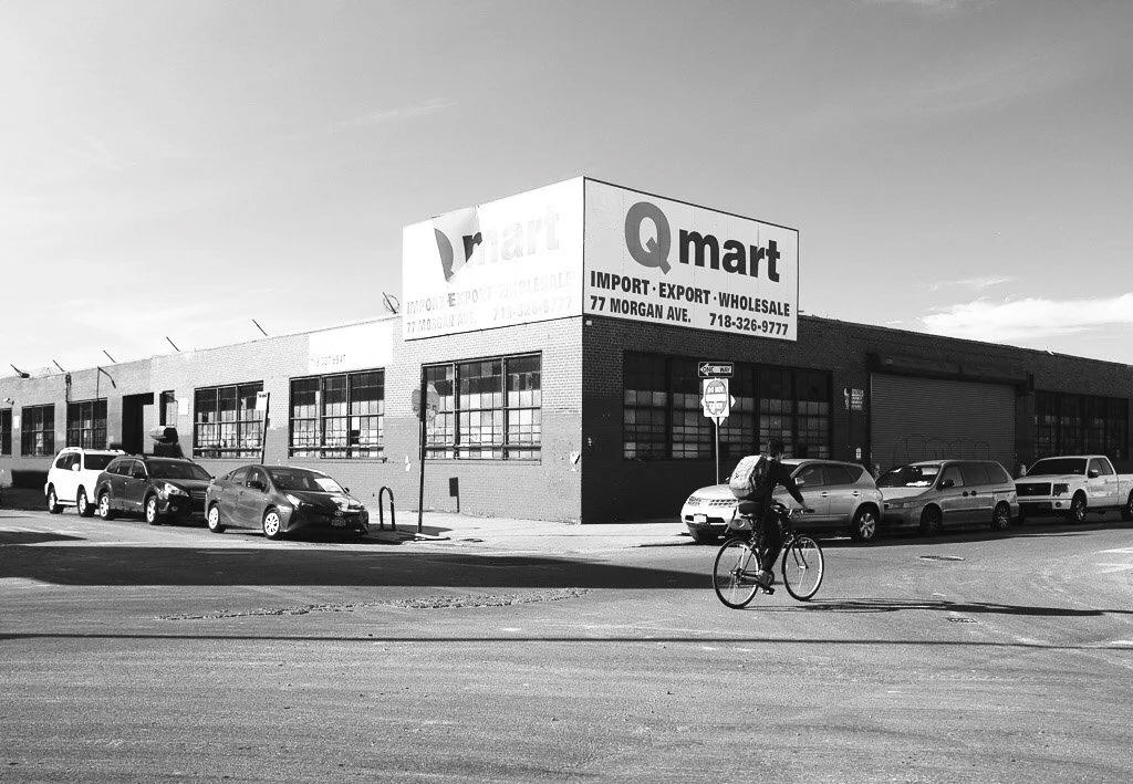A black and white photo of a commercial building with a sign that reads 'Qmart' and advertises import, export, and wholesale goods. There are several parked cars along the street and a person riding a bicycle in the foreground.
