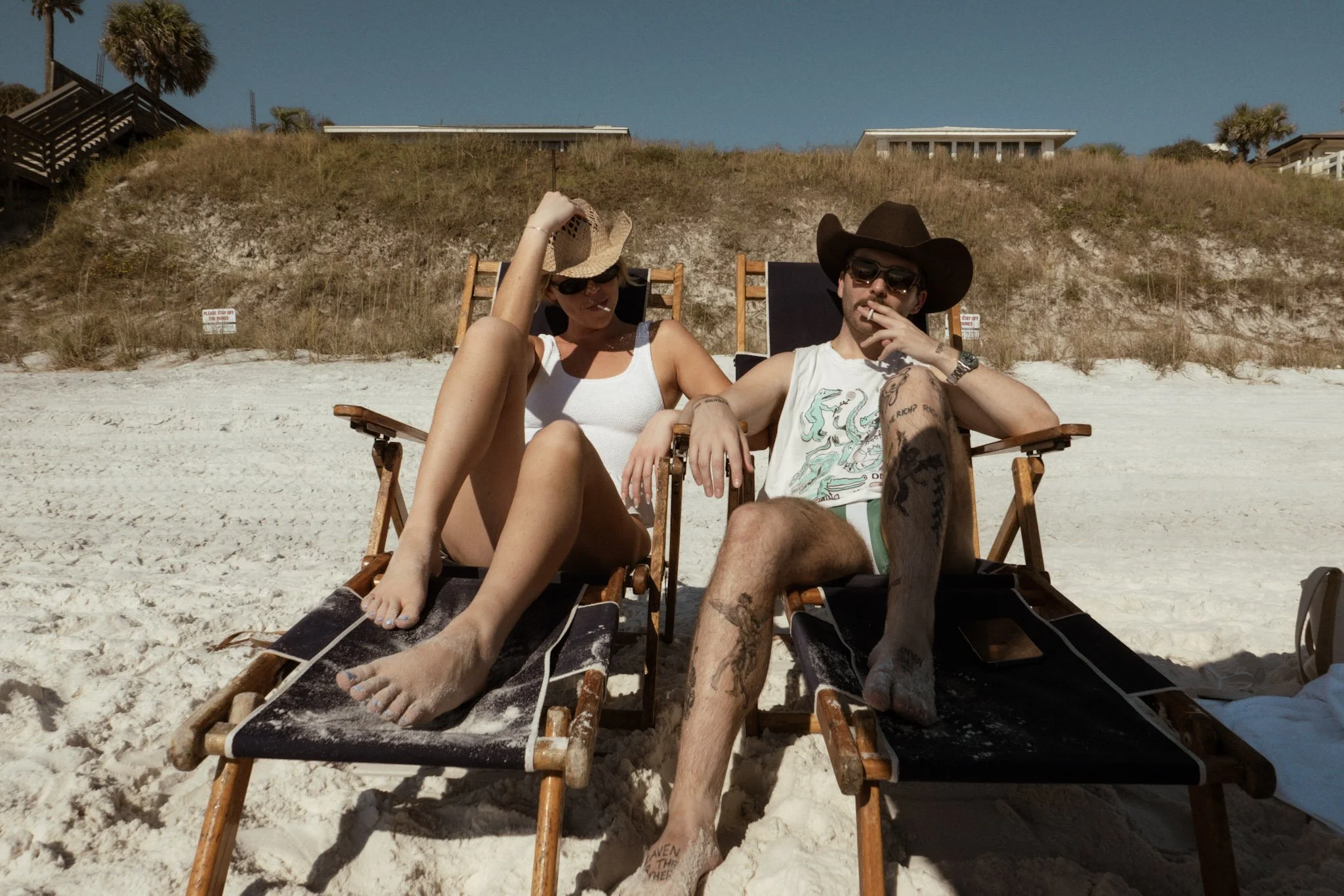 A man and woman relaxing on beach chairs on a sandy beach, wearing sunglasses and hats, with a grassy dune and houses in the background.