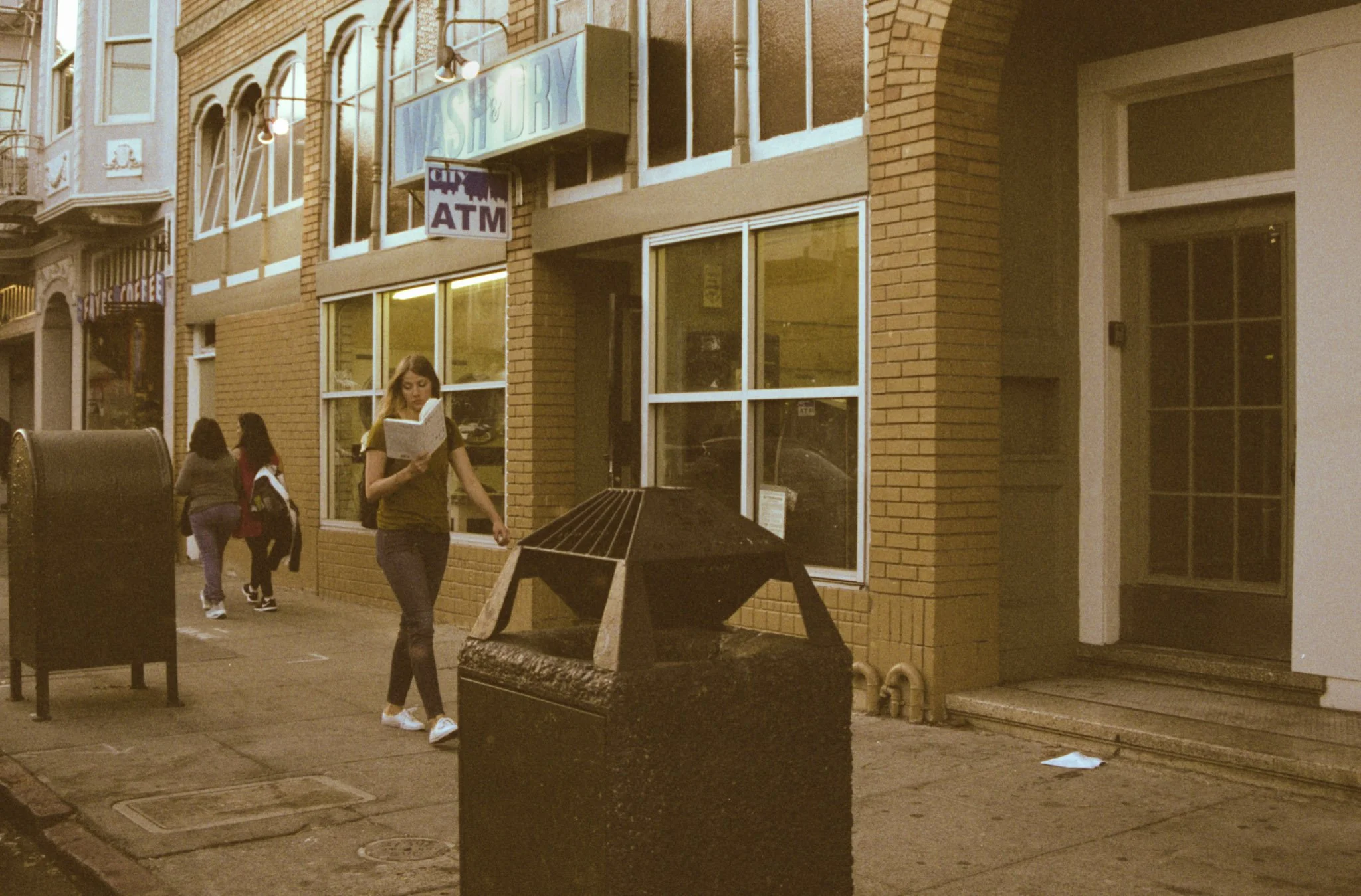 City street scene at dusk showing a brick building with large windows, a woman reading a book walking, and a sign indicating an ATM.
