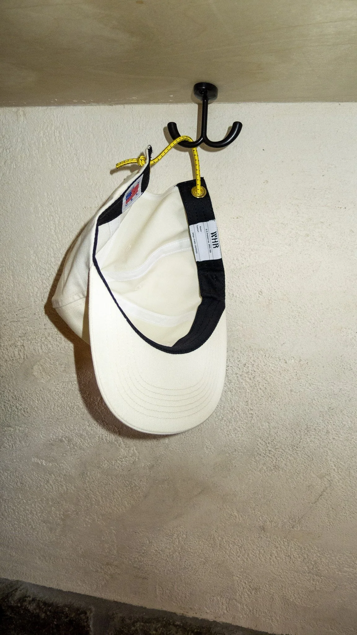 A white baseball cap hanging on a black hook, with a yellow measuring tape attached to the hook, on a textured beige wall.