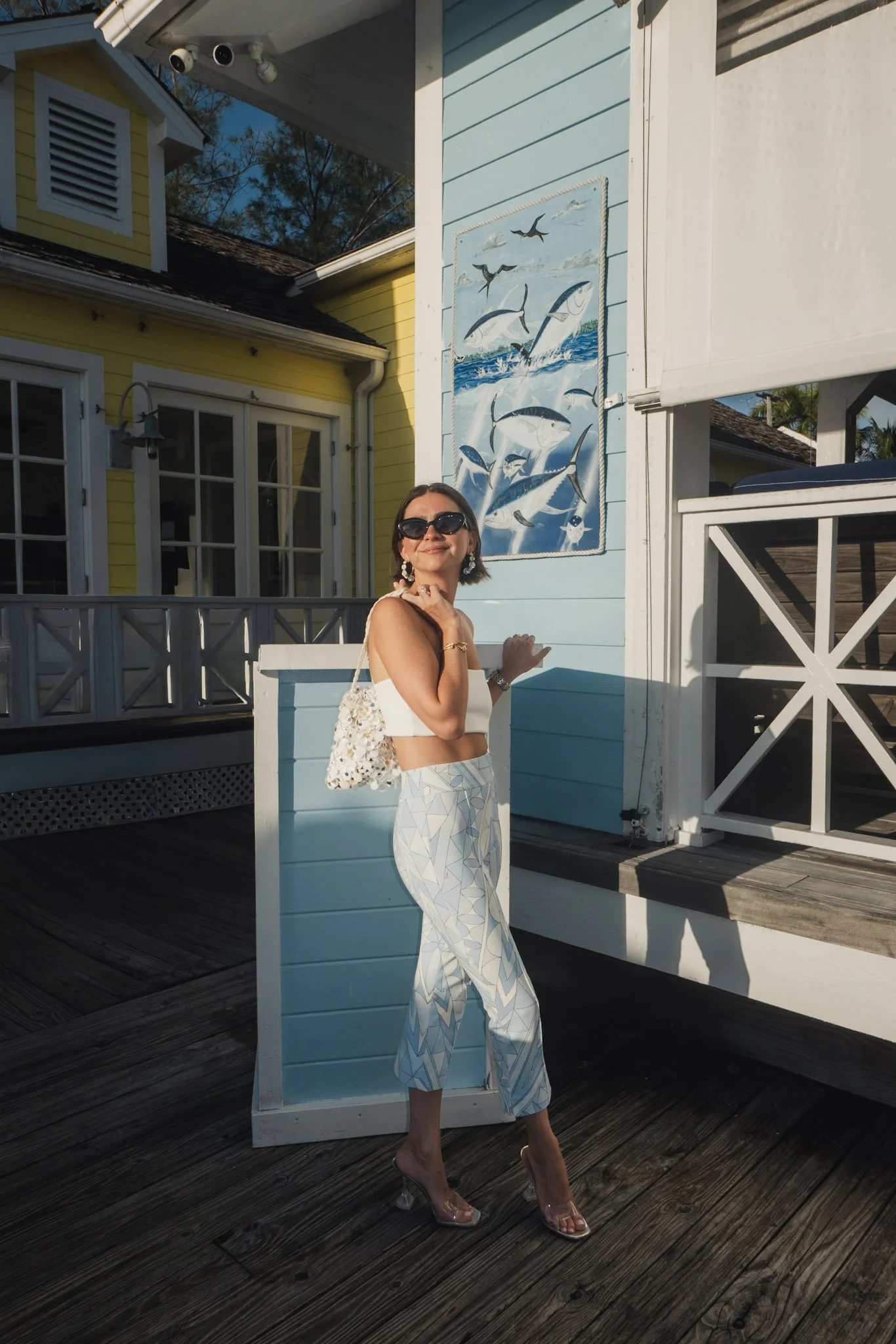 Woman standing outside on a wooden deck, smiling and wearing sunglasses, a white crop top, and white patterned pants, near a painted blue wall with a mural of jumping fish and flying birds.