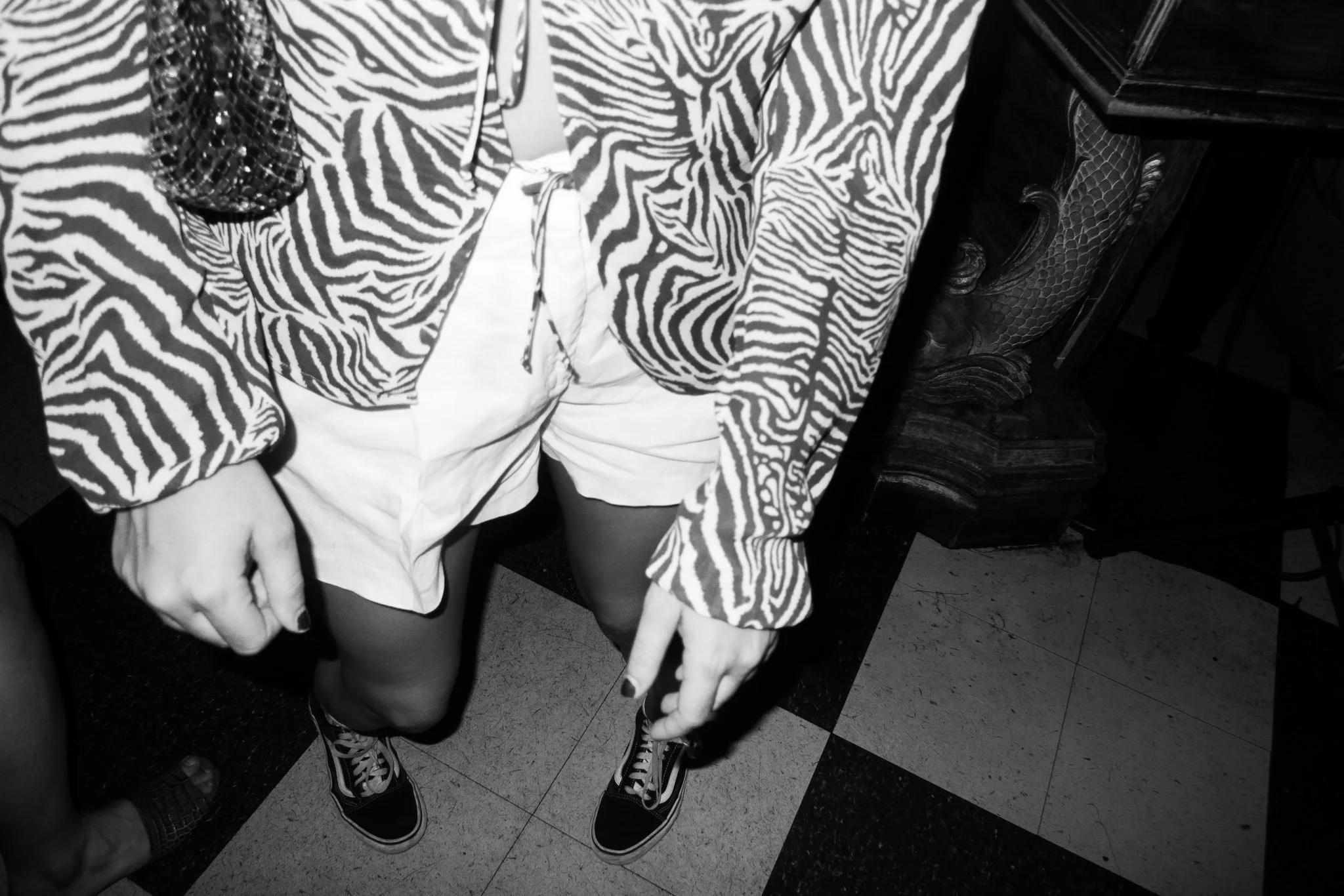 Two people standing close together, dressed in matching zebra print clothing, with one person pointing downwards, standing on a black and white tiled floor, in a room with black furniture.