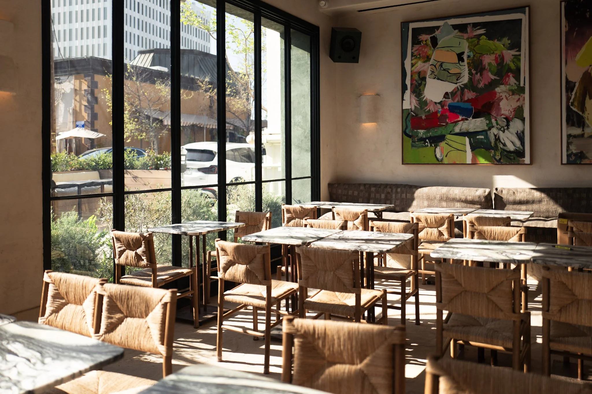 Interior of a restaurant with large windows, wooden chairs with woven seats, and abstract paintings on the wall.