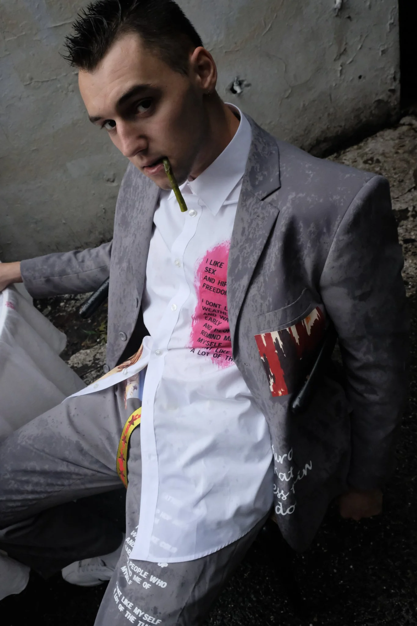 A man sitting outdoors against a concrete wall, wearing a gray suit and white shirt with pink and yellow printed text, holding a lit joint in his mouth. The man has dark, spiked hair and a serious expression.