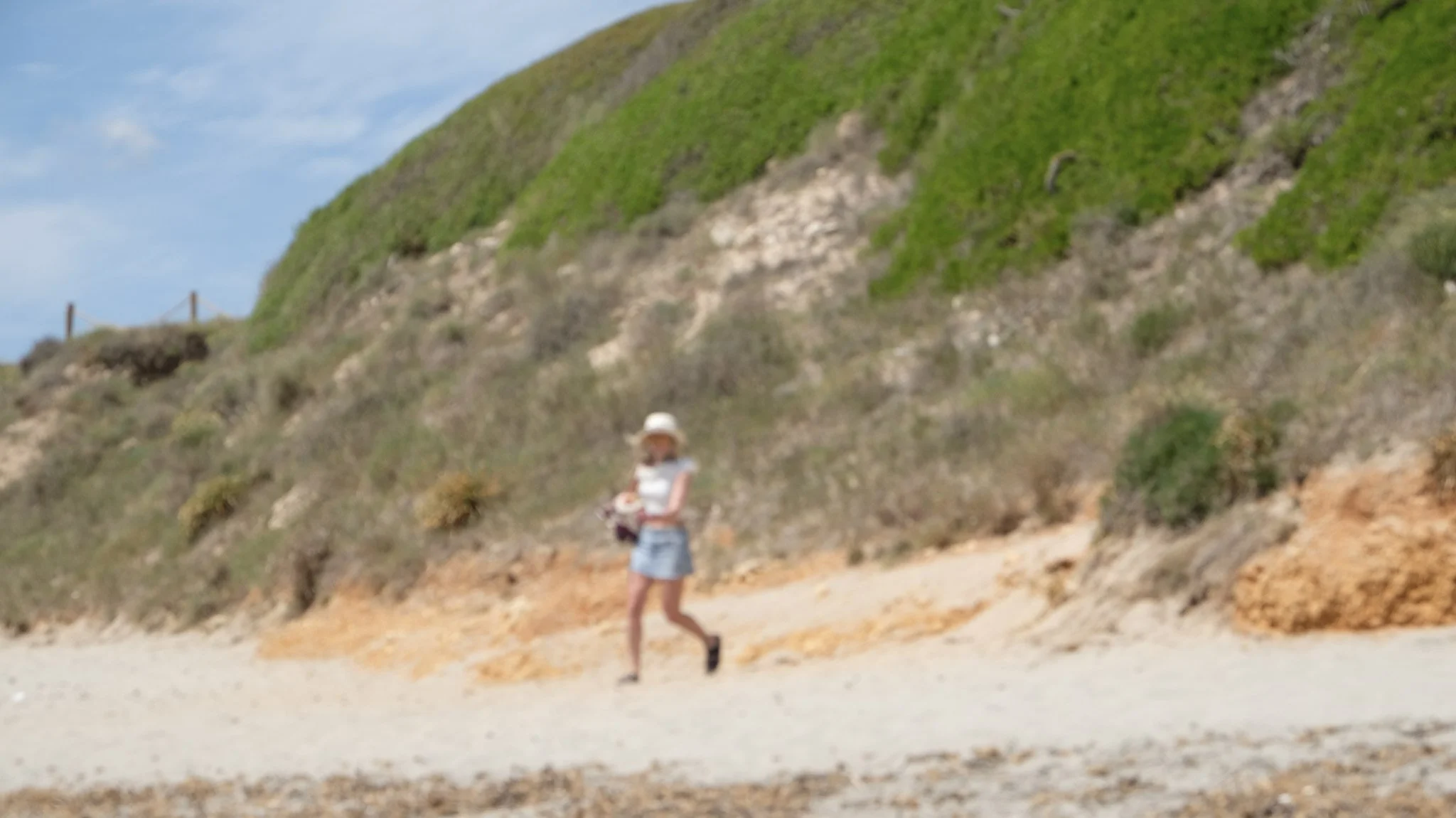 A person walking along a sandy beach with green hillside and blue sky in the background.