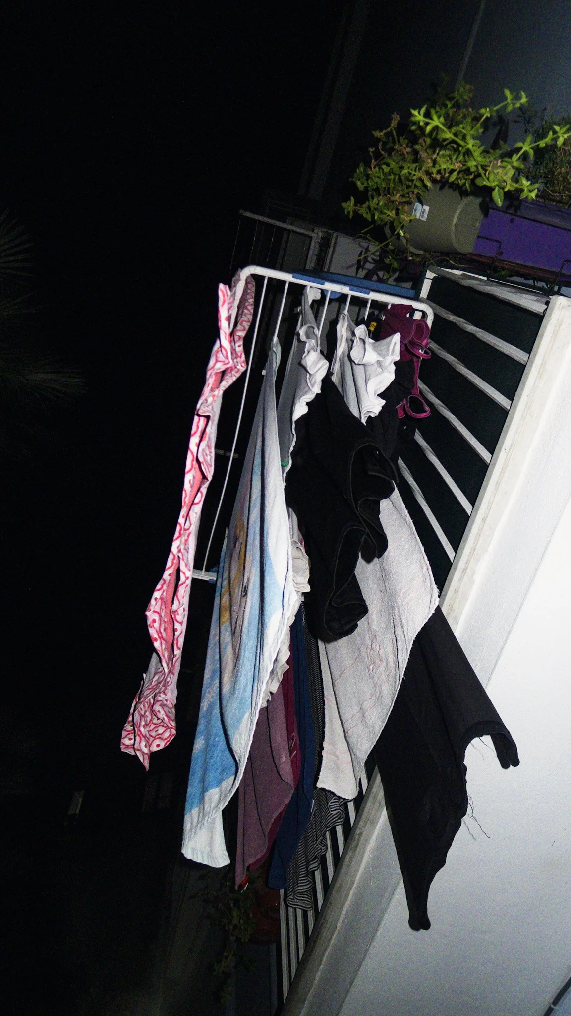 Clothes hanging on a balcony railing at night, with a potted plant and a dark sky in the background.