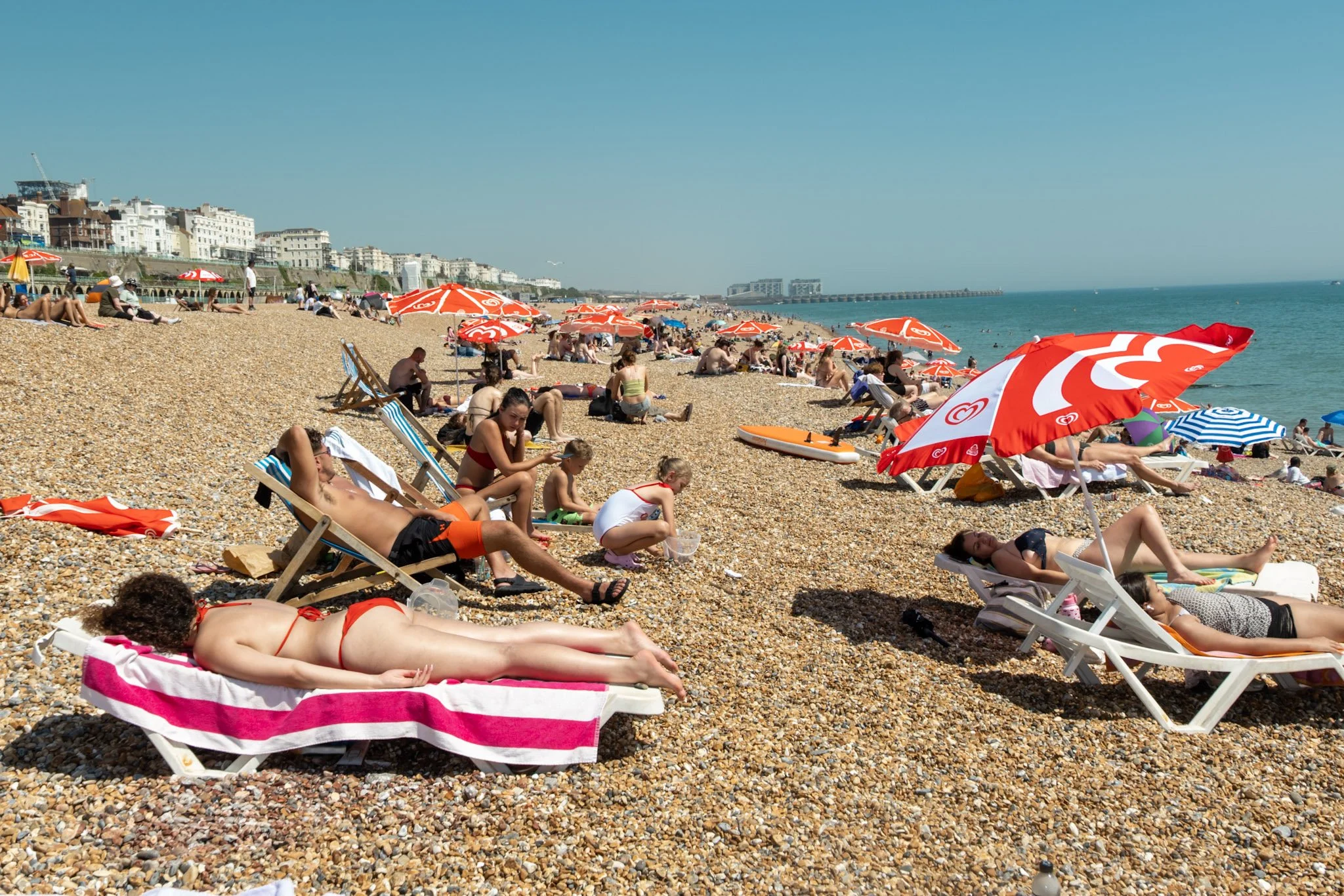 People sunbathing and relaxing on a pebble beach with umbrellas, in front of buildings along the shoreline, under a clear blue sky.