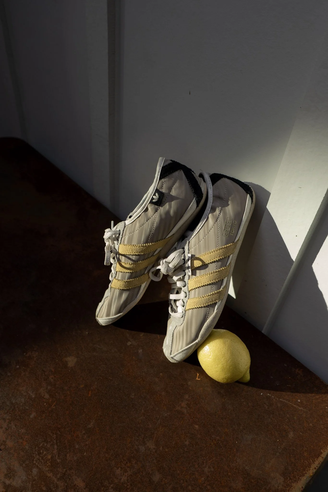 A pair of beige sneakers with yellow stripes resting against a white cabinet, with a yellow lemon on the wooden surface nearby.