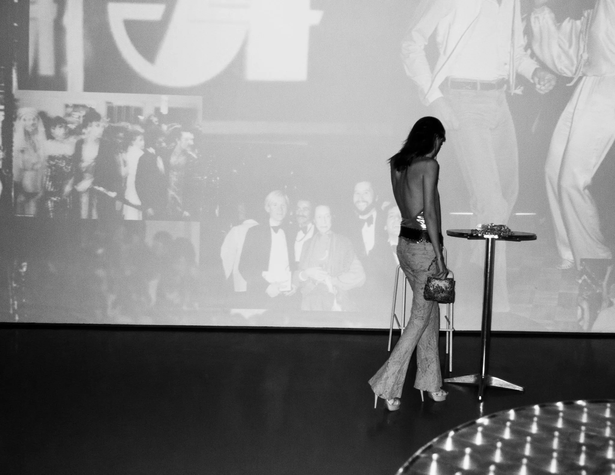 Black and white photo of a woman standing near a table with a decorative object, with a large screen or wall behind her displaying images of smiling people in formal attire and a large letter 'E'.