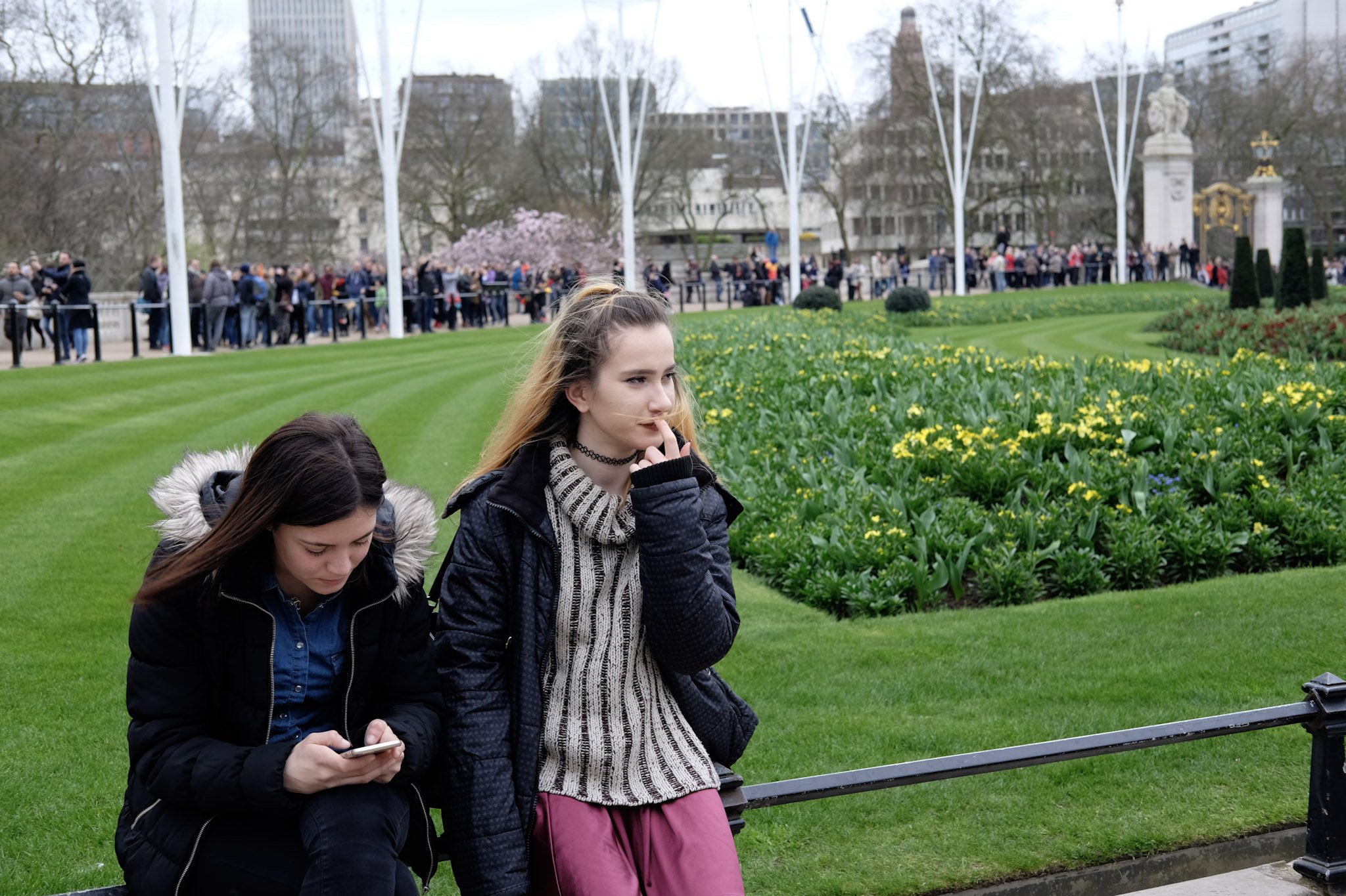 Two young women sitting on a park bench; one is looking at her phone, and the other is touching her nose, with a large crowd in the background near a fountain in a city park.
