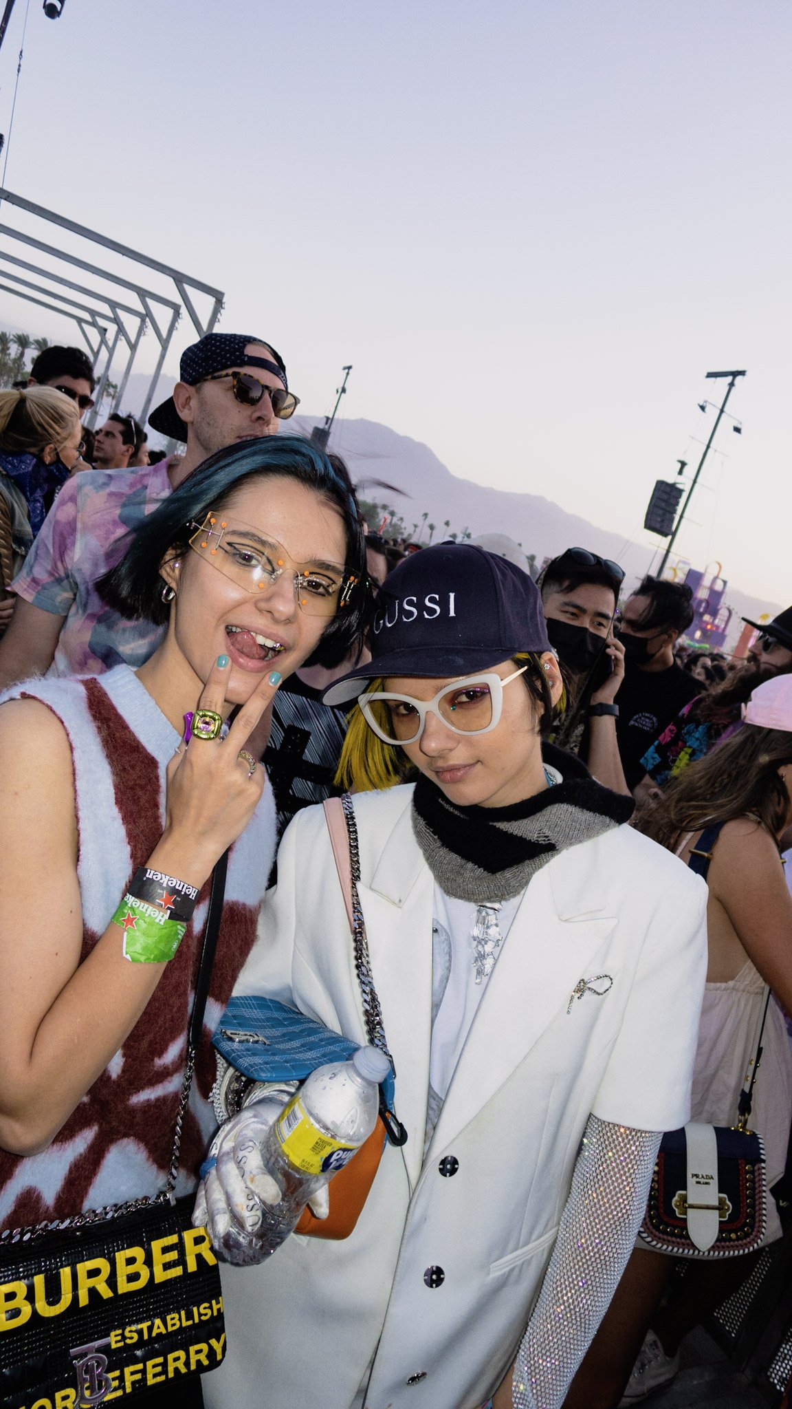 Two young women posing at an outdoor event with a crowd behind them. One has blue hair and glasses with decorations, making a peace sign, and the other has yellow hair, white glasses, and a white blazer.