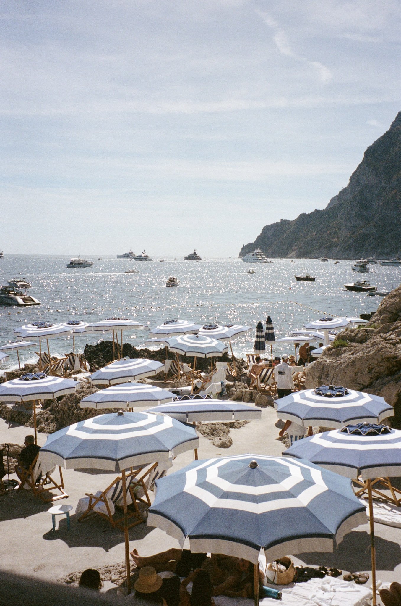 Beach scene with numerous white and blue striped umbrellas, wooden chairs, and people relaxing on the sand. The background features a calm sea with several boats and yachts, and a large rocky hill on the right side.