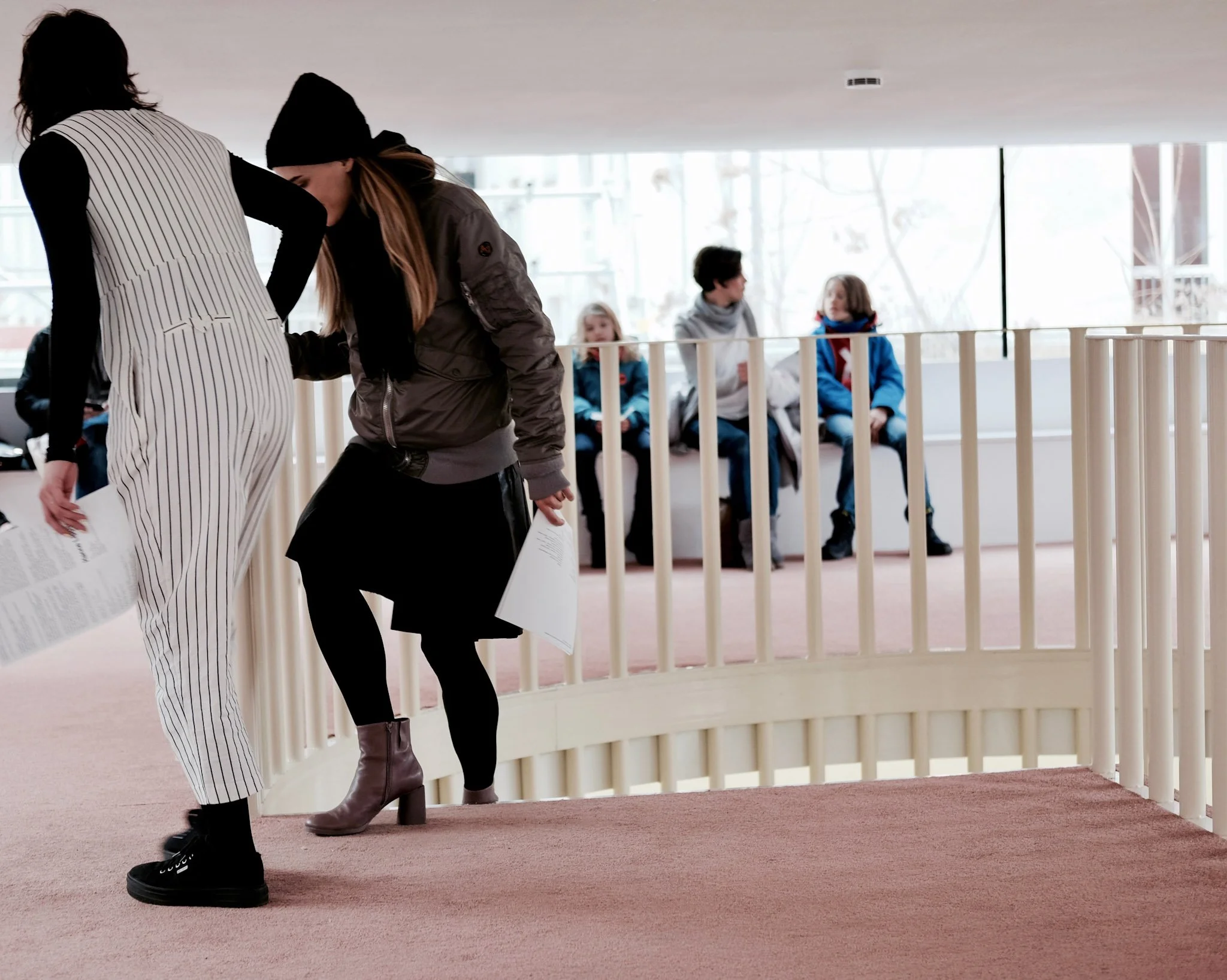 People walking through an indoor space with large windows, with children sitting on benches behind a white railing in the background.
