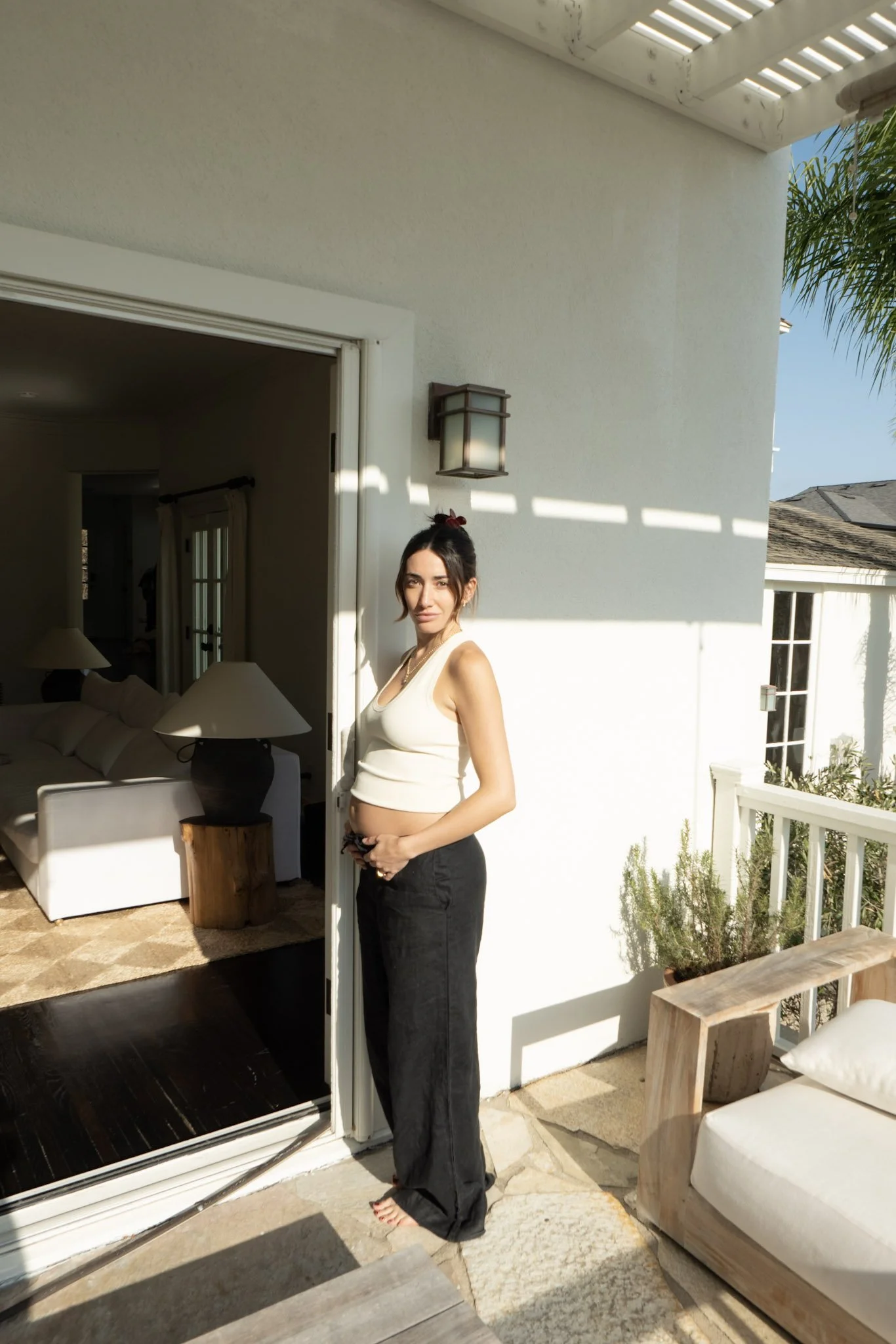 A young woman with dark hair tied in a bun, wearing a white tank top and black pants, standing on a balcony next to a sliding glass door, holding her pregnant belly, with sunlight casting shadows on the white wall behind her.