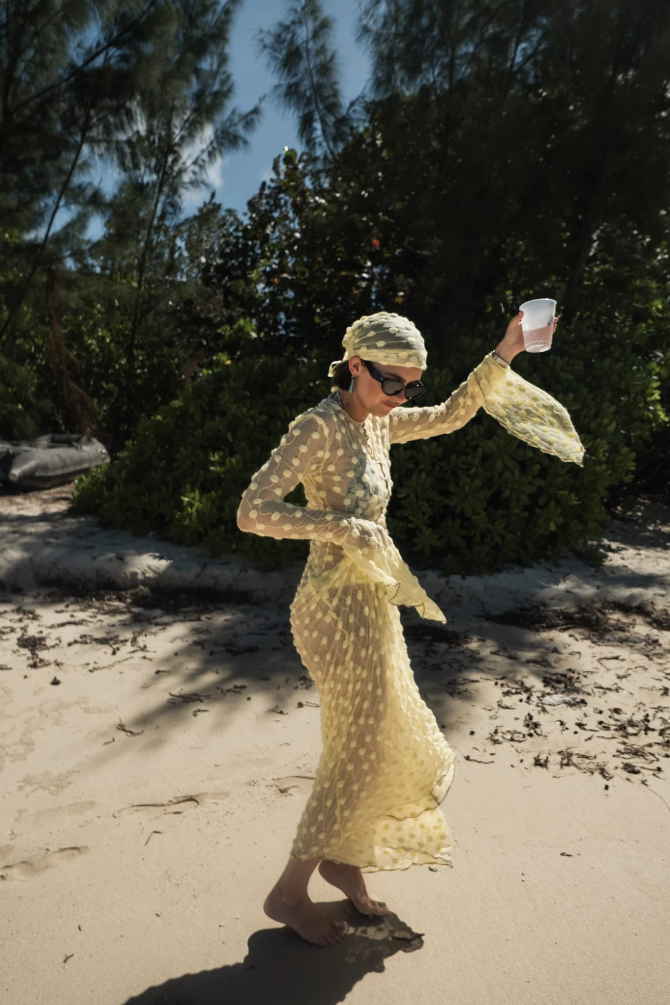 A woman in a yellow dress with polka dots, sunglasses, and a headscarf is walking barefoot on the beach, holding a plastic cup, with trees and blue sky in the background.