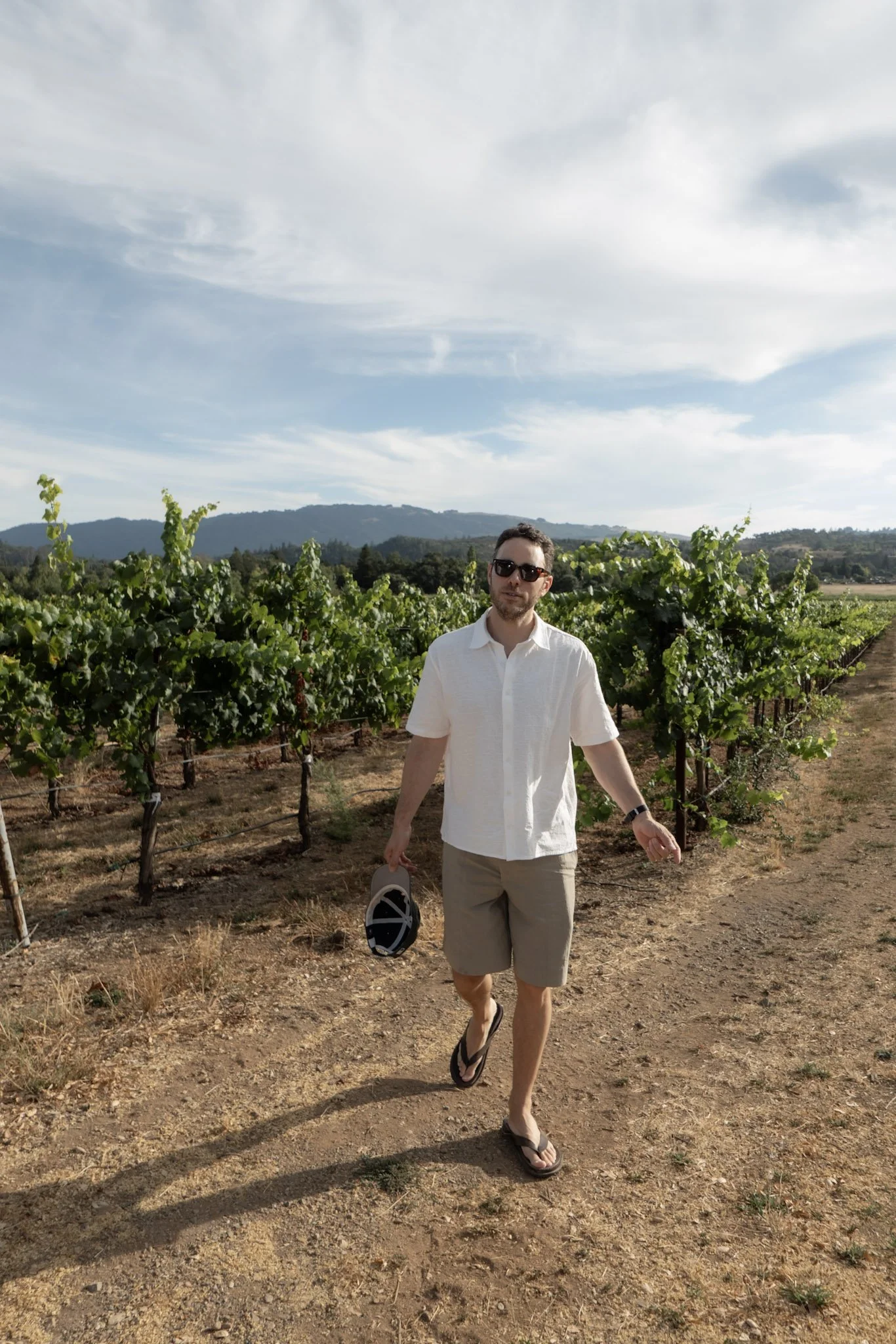 A man walking through a vineyard on a sunny day, holding a hat and wearing sunglasses, a white shirt, khaki shorts, and flip-flops.