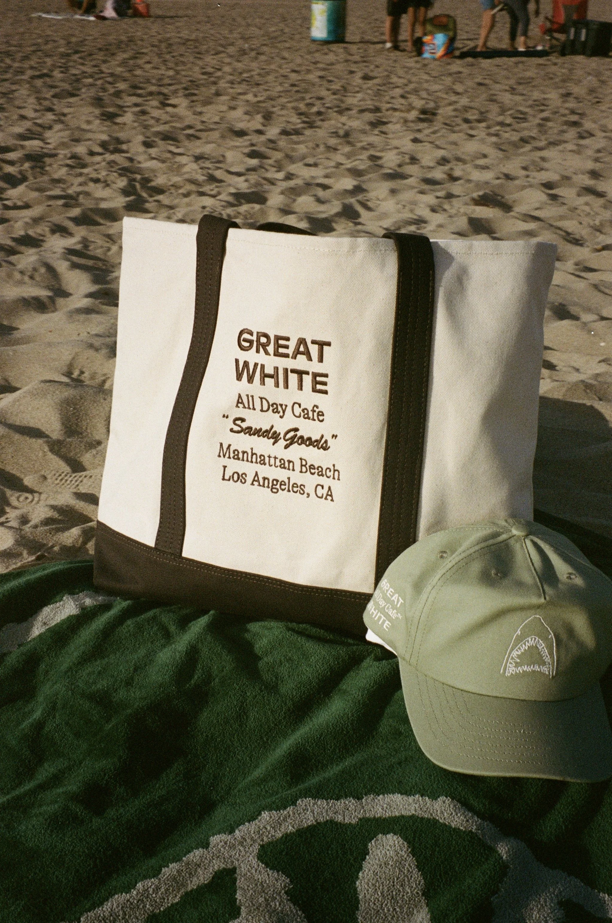 Beach scene with a white tote bag that has text about Great White All Day Cafe, a tan bucket hat, and a green towel or blanket on the sand. In the background, there are people and beach equipment.