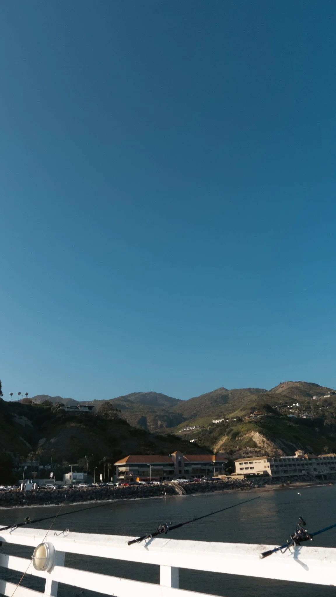 View of coastal area with hills, buildings, and parking lot near the water, as seen from a boat with fishing rods.